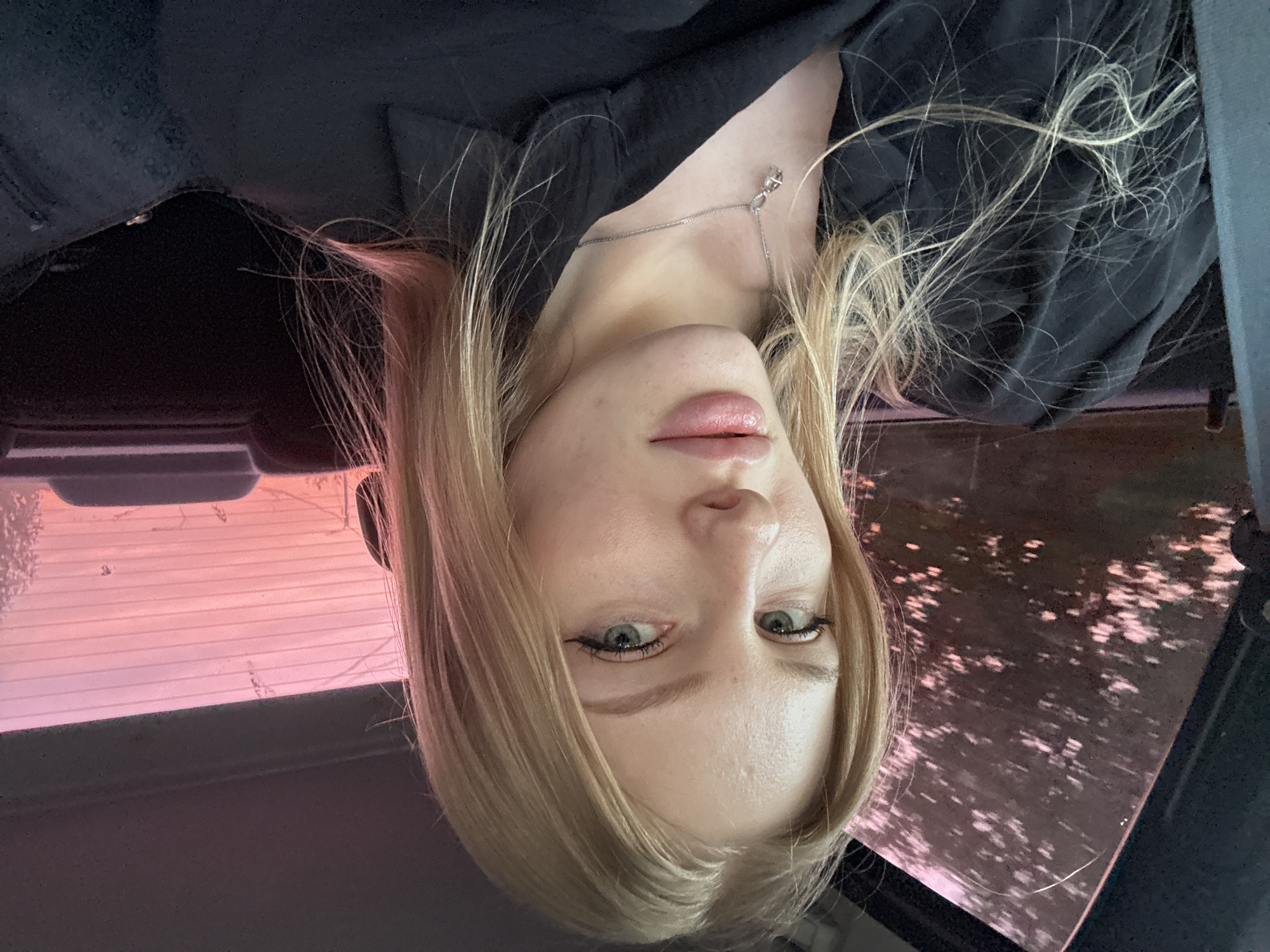 A woman with blonde hair and green eyes is pictured inside a vehicle, likely a car, in Kondratovo, Russia. The image is inverted. She is wearing a black shirt or jacket and a silver necklace. The exterior visible through the tinted car windows has a pink hue, possibly indicating a sunset or a reflection of pink light. There are indistinct objects or scenes visible outside.