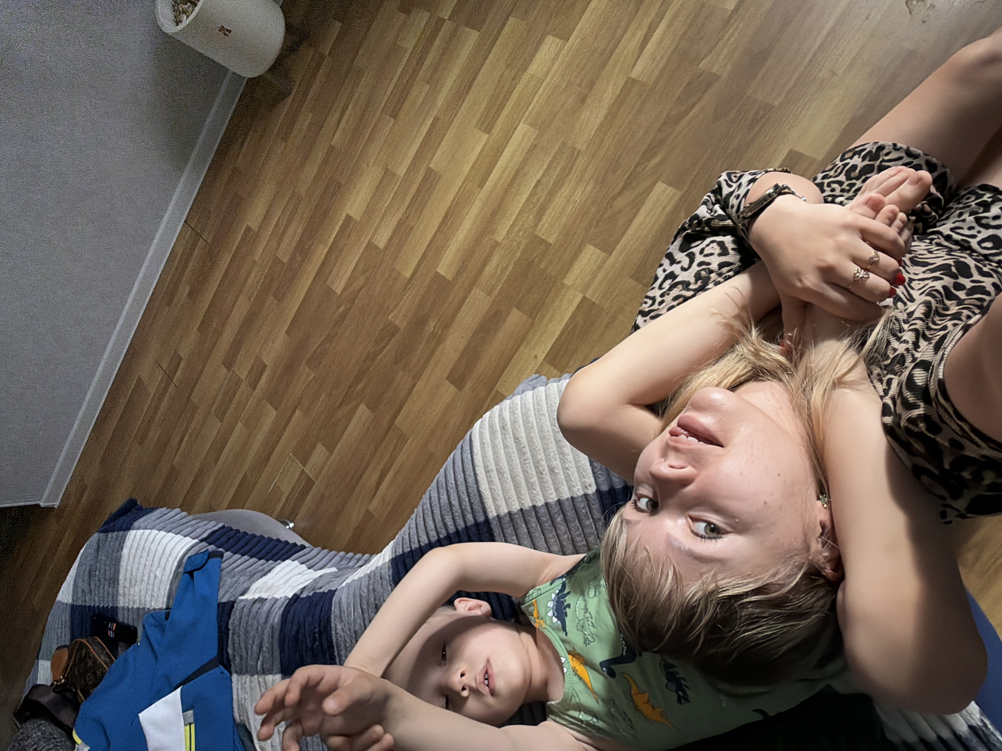 A woman and child are lying on a gray and blue striped sofa or bed in what appears to be an indoor location. The woman is wearing a leopard-print dress and is lying on her back with her arms crossed, looking at the camera. She has blond hair and is wearing jewelry on her hands and wrists. The child is lying beside her, wearing a green dinosaur-patterned shirt, also looking at the camera. A blue and white jacket and a small brown purse are on the left side of the frame. The floor is wooden, and a white planter is in the upper left corner. The location may be in an apartment in the city of Chaikovsky, Russia.
