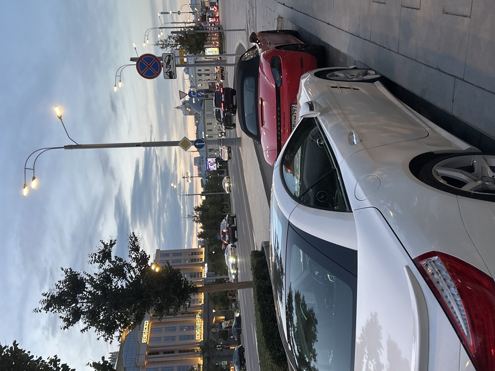 The image depicts a street scene in a city at dusk, with cars parked along the sidewalk and traffic on the road. In the foreground, two convertible sports cars are parked: a white car on the left and a red Porsche on the right. The white car is partially visible, showing its rear end and side profile. The red Porsche is a Porsche 718 Boxster S, as indicated by the badging. Beyond the parked cars, the street is busy with moving traffic. Several other vehicles, including SUVs and sedans, are visible on the road.
The background features a large, modern building with illuminated windows and signage that reads "Бизнес Центр" (Business Center). Streetlights line the sidewalk, their lamps illuminated, casting a warm glow. Traffic signs, including a "No Parking" sign and a towing warning sign, are also visible. The sky is overcast with hints of sunset colors. Trees with dark foliage are scattered along the street and sidewalk. The overall impression is that of an urban environment in the evening, with a focus on the parked luxury vehicles.