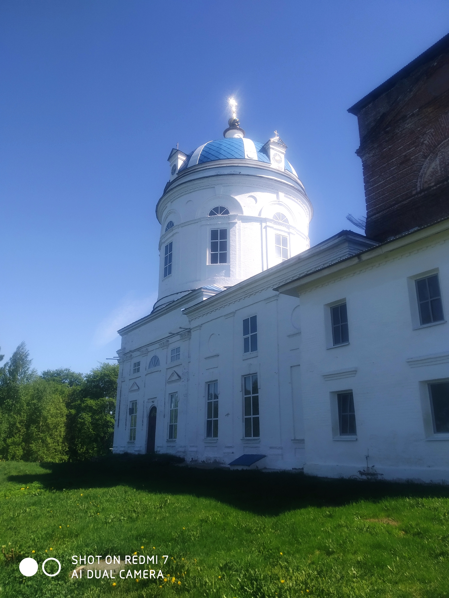 The image shows a white building with a blue-domed tower located in Ochansk, Russia. The building has multiple windows and is surrounded by green grass and trees. The sky is clear and blue. The tower is topped with a cross. Part of a brick building is visible on the right side of the frame.