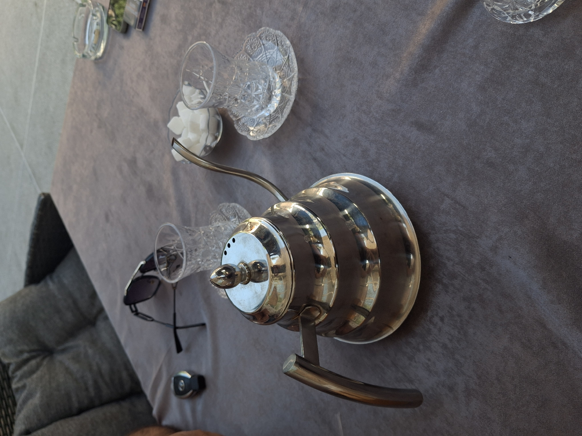 The image shows a table covered with a grey tablecloth. On the table, there are several objects including a silver kettle with a gooseneck spout, two clear glass cups with ornate patterns, a small bowl containing white sugar cubes, a pair of sunglasses, and a small black object that appears to be a remote control or key fob. A portion of a grey upholstered chair is visible in the lower left corner, suggesting the setting is an indoor or covered outdoor seating area. The lighting in the image suggests it is daytime, and the overall atmosphere appears relaxed and domestic. There are no visible people or text in the image. The scene might be set up for a coffee or tea break. The location context provided is Rostov-on-Don, Russia.