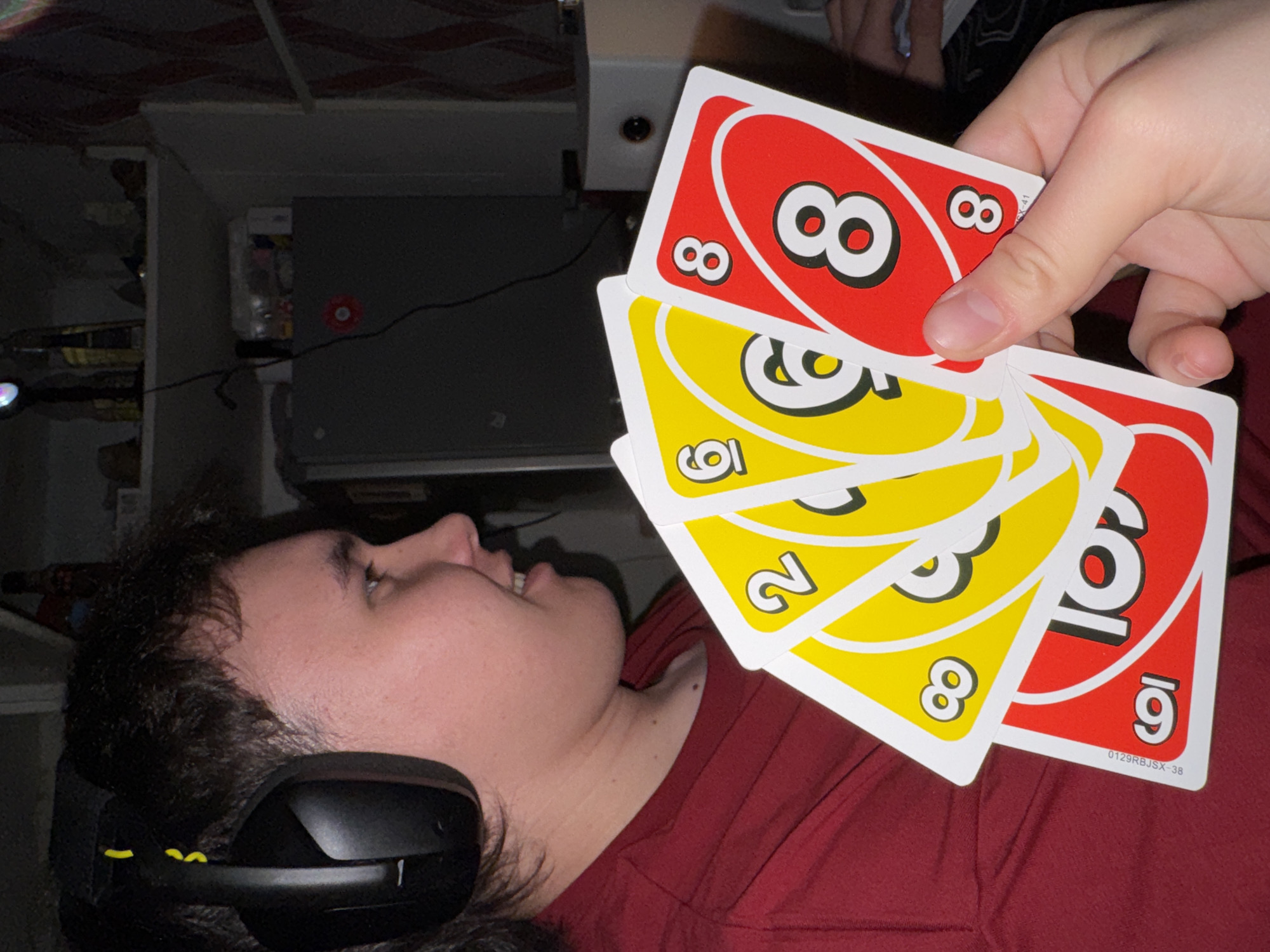A person is holding a hand of Uno cards in front of another person who is wearing a headset. The cards are primarily red and yellow. The person holding the cards is facing downward towards the other person. The second person is wearing a red shirt. The location cannot be determined.