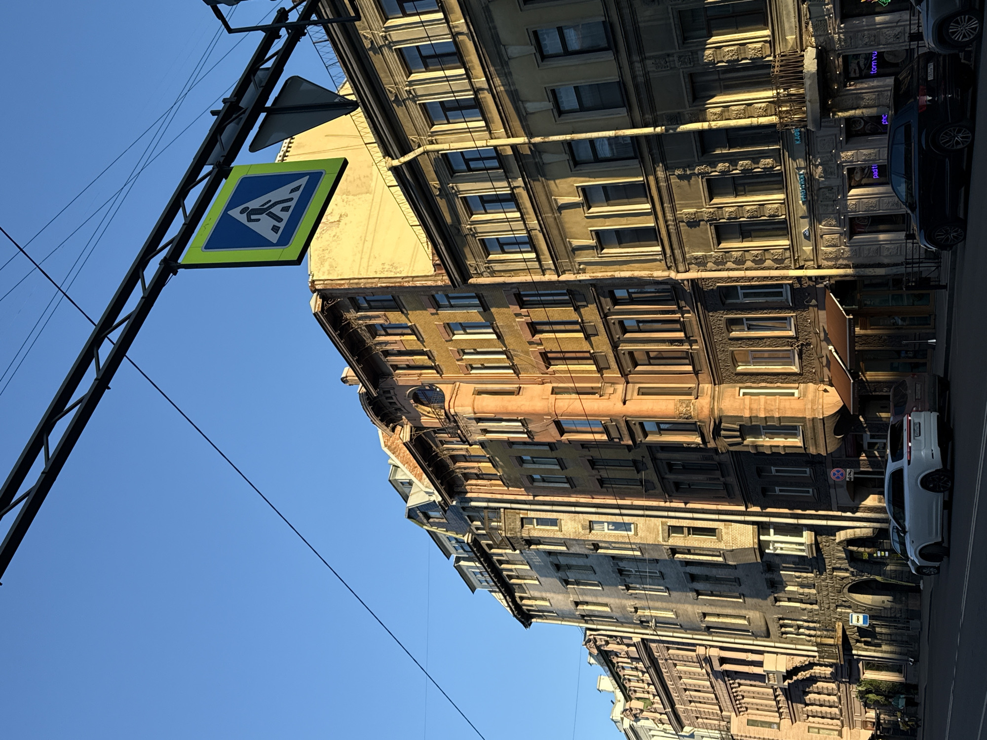 The image displays an urban street scene in Saint Petersburg, Russia, characterized by grand, multi-story historic buildings under a clear, bright blue sky. The perspective is tilted, looking upwards from street level.
On the right, a row of ornate buildings dominates the frame. These structures feature intricate architectural details such as decorative window frames, cornices, and textured facades in shades of grey, beige, and light brown. Sunlight illuminates the building fronts, creating distinct shadows that suggest it is daytime, possibly morning or late afternoon. Visible drainpipes run down the side of some buildings.
Overhead, a black metal gantry supports a prominent pedestrian crossing sign. This sign is a blue square with a white pedestrian figure on a diagonal zebra crossing, all enclosed within a distinctive green-yellow border. Several dark wires stretch across the clear sky from this structure.
At street level, a paved road hosts several parked cars, including a dark-colored SUV/crossover and a white SUV/crossover. A license plate visible on one of the dark cars shows the number "78," a regional code for Saint Petersburg. Other visible street signs include a circular red no-parking/stopping sign and a blue square parking sign with a white 'P'. Some commercial establishments on the ground floors of the buildings display partially visible neon signs with text such as "tom yu" and "pattı". No people are visible, and the scene depicts a quiet moment on a historic city street with clear, sunny weather.