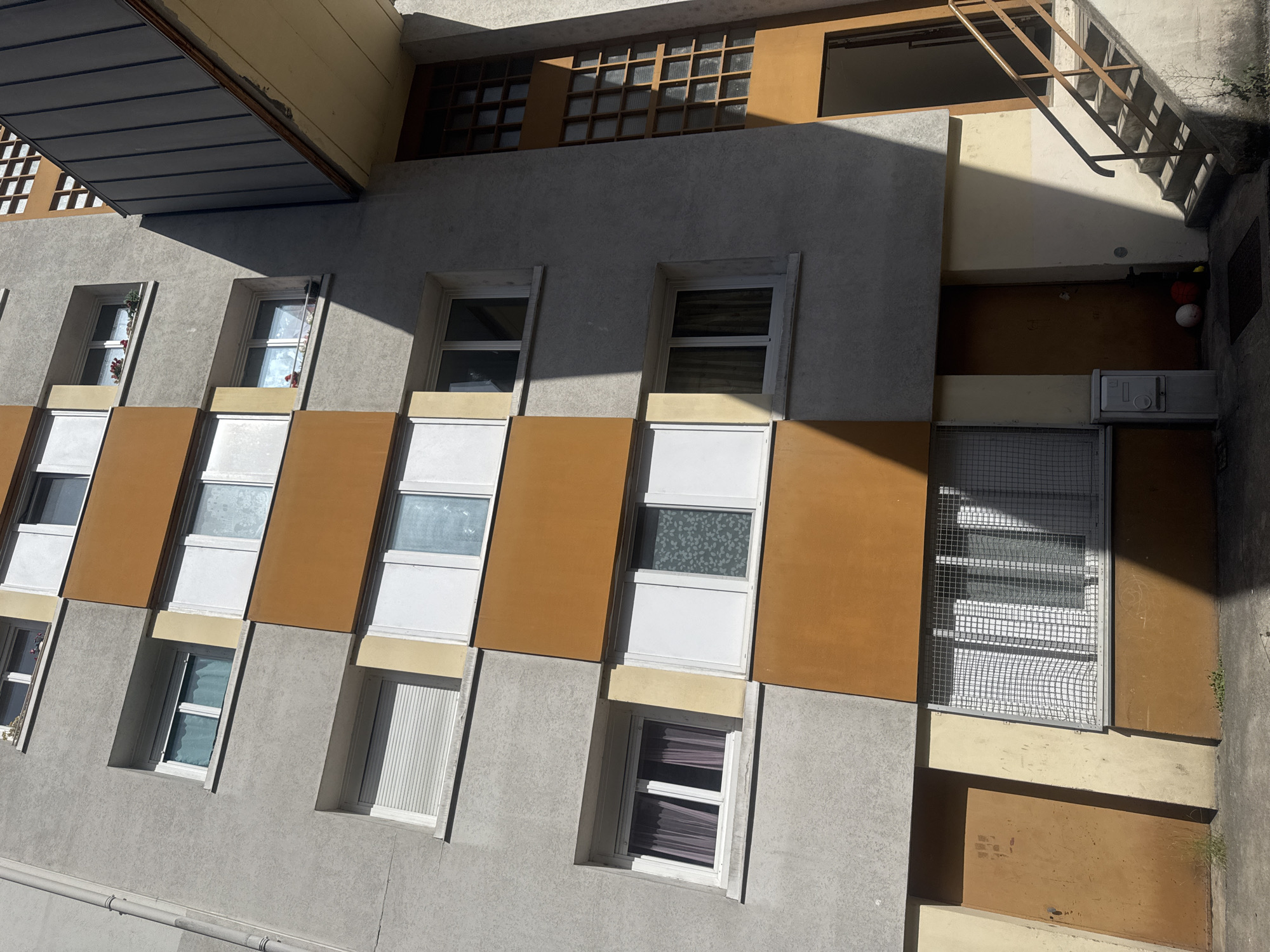 This is a full exterior, eye-level, medium shot of a building in Saint-Claude, France, likely an apartment building or a public housing complex. The building has a facade of gray concrete interspersed with horizontal bands of orange and cream-colored panels. Multiple windows are visible, some with white frames and blinds, others with darker curtains or frosted glass. Some windows have small flower boxes outside. To the right, a dark, modern architectural element with a metal facade extends upwards and angles across the building. Below this, a recessed section of the building features a tall, grid-like window structure with wooden mullions, illuminated by sunlight. At the base of the building, two brown doors are visible. One on the left is slightly ajar, and the other on the right is open, revealing an entranceway. A set of concrete stairs with metal railings leads up to the open doorway on the right. A metal mailbox is situated near the lower left door. Sunlight casts strong shadows across the facade and the ground, indicating a bright day. No people are present in the image.