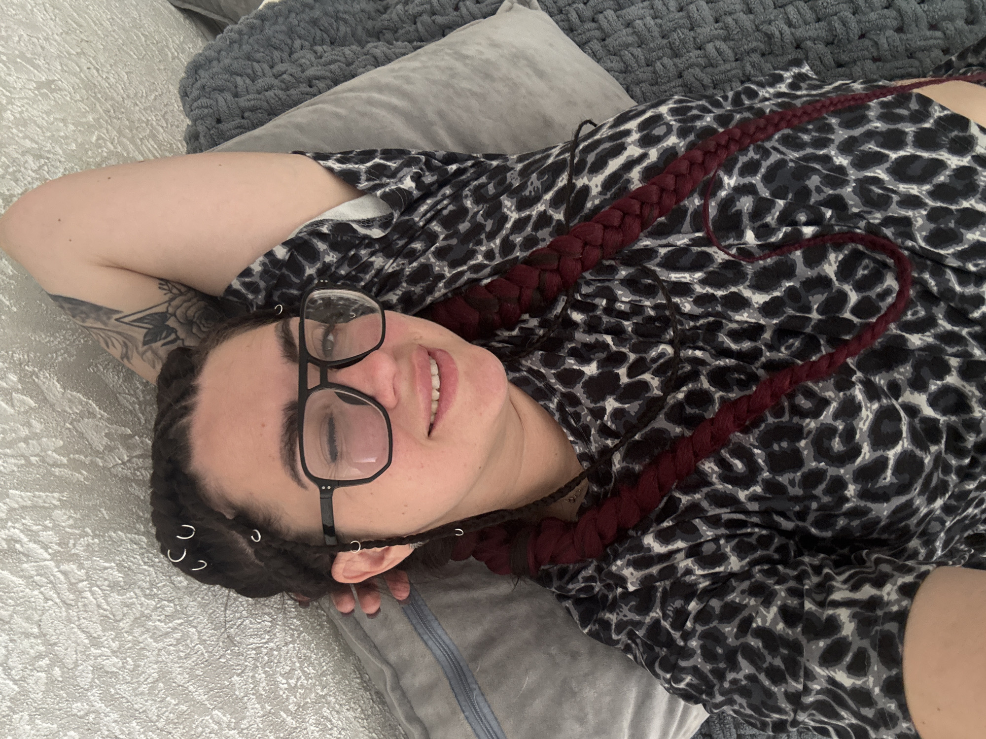 A young person with dark braided hair, adorned with several silver hoops, is lying on a bed with their head propped on a pillow. They are wearing glasses and a black and white leopard-print top. Their eyes are closed, and they have a slight smile. A tattoo is visible on their arm, and their hair is styled in two thick braids. The background is a textured wall.