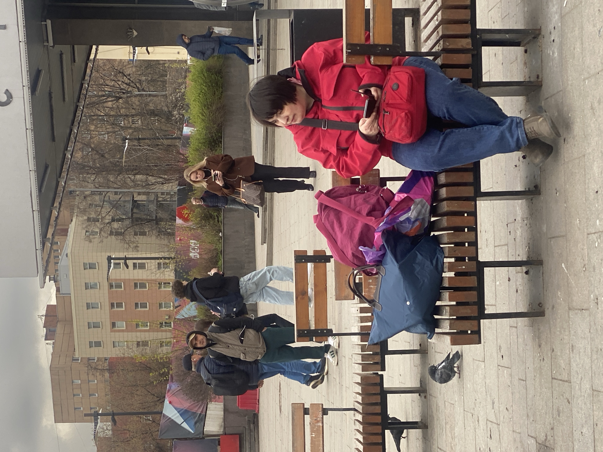 The image captures an urban scene, likely in Moscow, Russia, considering the architectural style and general ambiance. A woman wearing a red jacket and blue jeans is seated on a wooden bench, using her phone. Next to her is a pink backpack and a blue tote bag. Several other people are walking in the background, including a woman with a brown jacket and a man with a blue jacket.
Further in the background are buildings of apartments and office buildings. A few pigeons can be seen in the image.