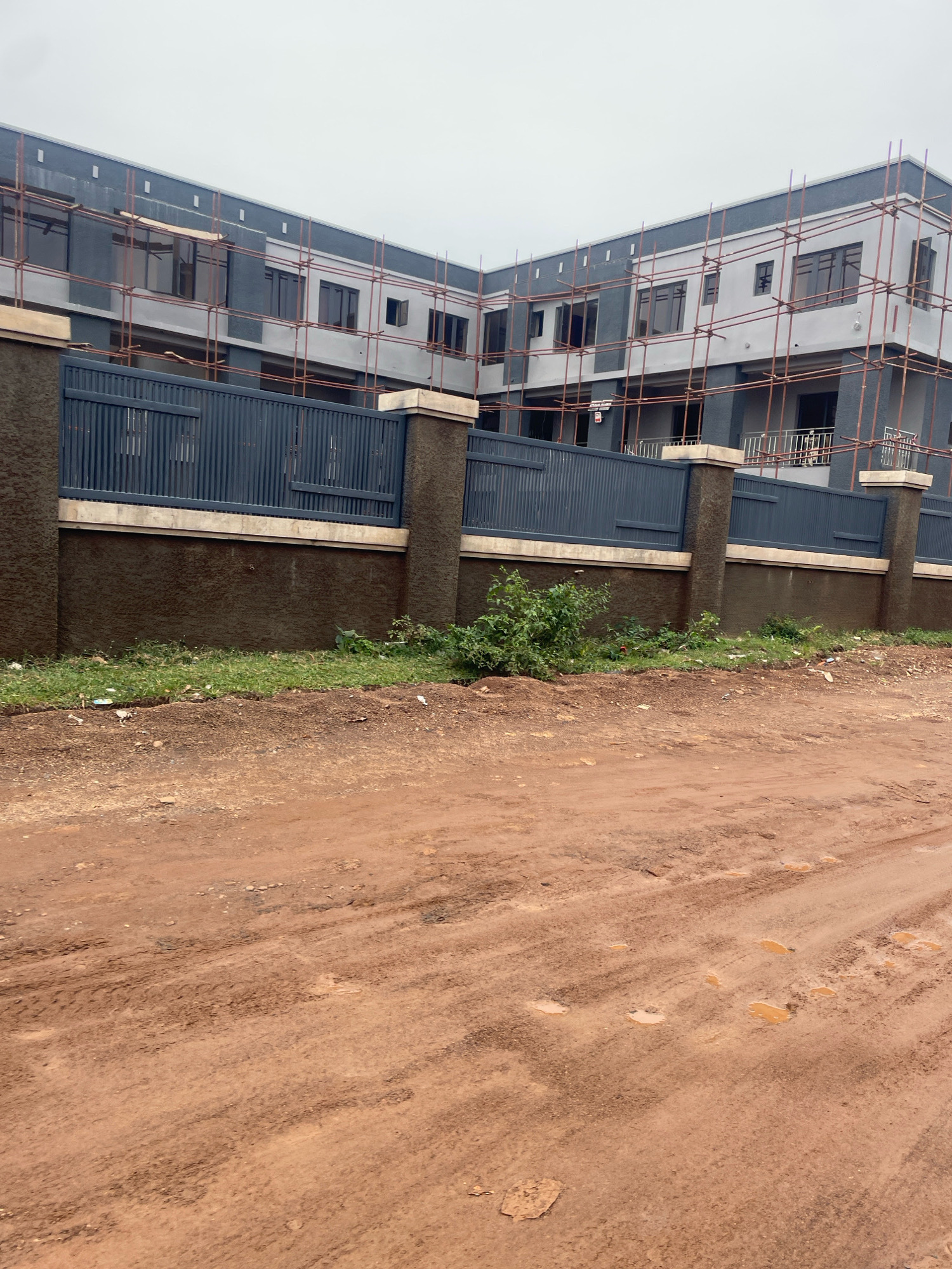The image depicts a large, multi-story building under construction in Abuja, Nigeria. The building is a modern design with a gray and dark gray exterior, featuring numerous windows. Scaffolding surrounds the upper levels, indicating ongoing construction work. In front of the building is a dark gray metal fence with vertical slats, supported by stout, dark brown pillars. A textured, dark brown wall runs behind the fence. A patch of green grass and some small plants are visible between the wall and the fence. The foreground of the image shows a rough, reddish-brown dirt road, with tire tracks and several puddles of water, suggesting recent rain. The sky is overcast and a pale gray.