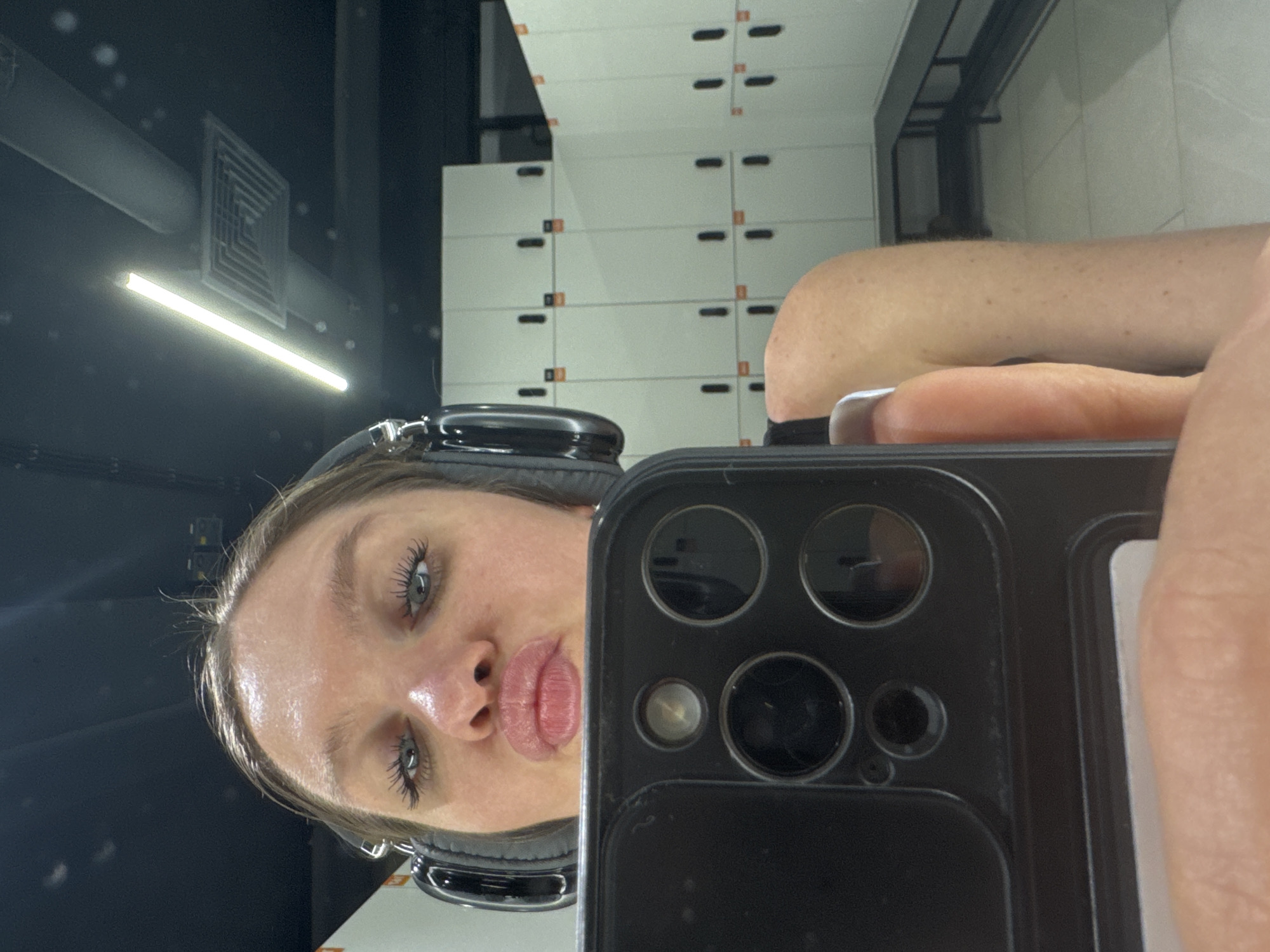 The image shows a woman taking a selfie in what appears to be a locker room. She is wearing headphones and holding a smartphone up to the mirror, partially obscuring her face. In the background, rows of white lockers are visible. Based on the style of the locker room and the lighting, it is likely located in a fitness center or gym. It is impossible to determine from the image if the location is in Moscow, Russia.
