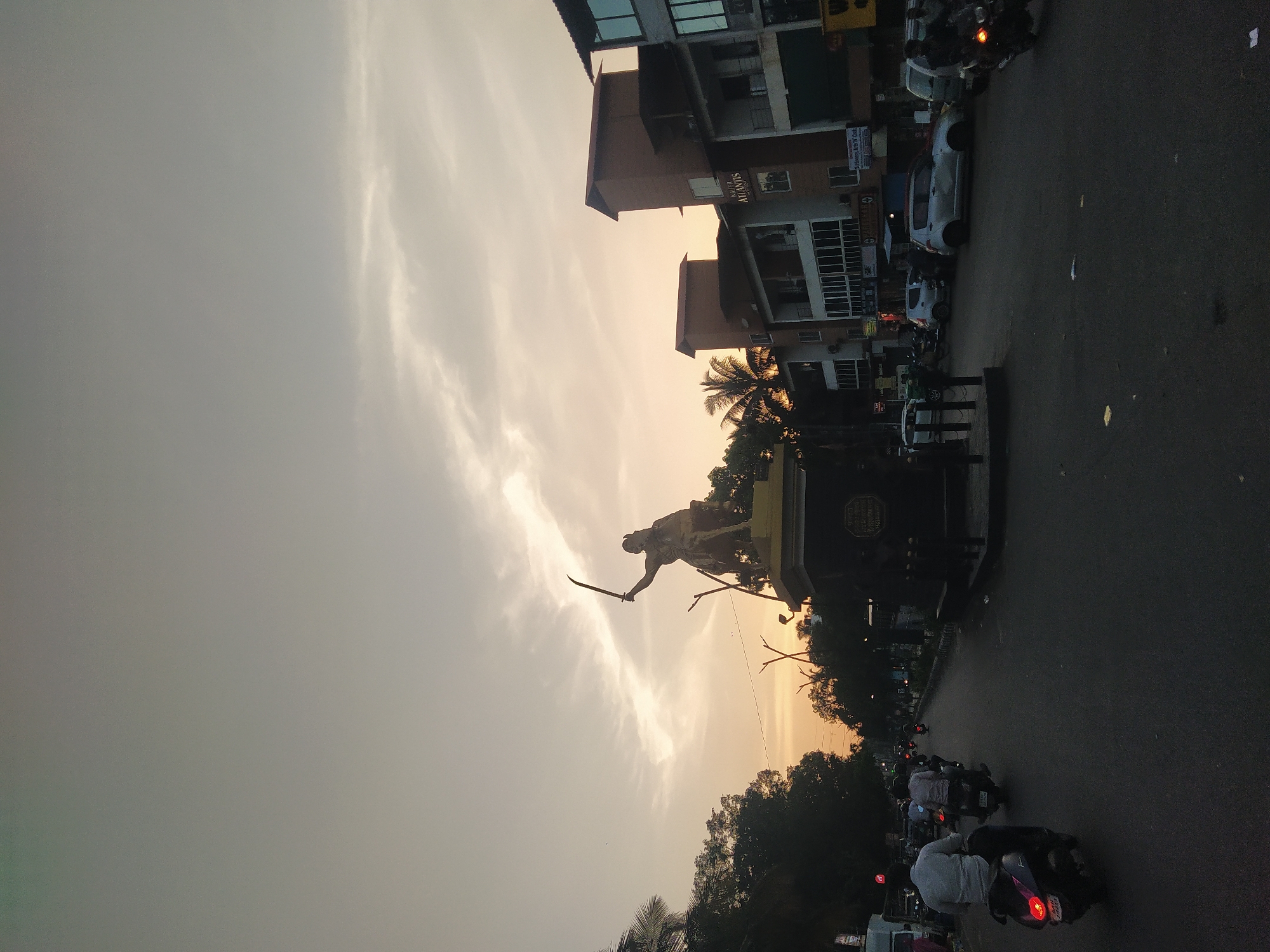 The image depicts a street scene in Oxel, India, at dusk or dawn. A statue of a man holding a sword is prominently featured in the middle ground. Behind the statue, buildings and trees are visible against a sky with a golden hue. In the foreground, the street is filled with motorbikes and scooters. Cars are parked or moving alongside the buildings. The photograph appears to be taken from an elevated position on the side of the road.