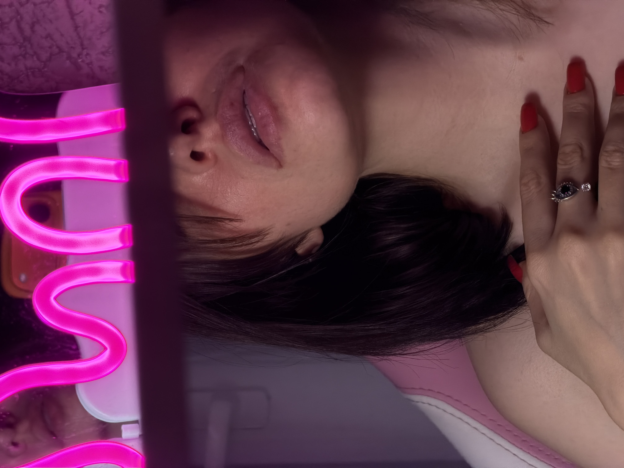 The image is a close-up shot of a young woman's face and upper chest, with a neon pink light fixture in the foreground. The woman has dark brown hair with bangs, and her skin appears smooth with a subtle pink blush on her cheeks. She is wearing red nail polish and a ring with a dark gemstone and a small clear stone on her left hand, which is resting on her chest. The background is dimly lit with purple and pink hues, suggesting an indoor environment, possibly a room decorated with neon lights. The overall mood is intimate and sensual. The image does not provide any information about the location within a city.