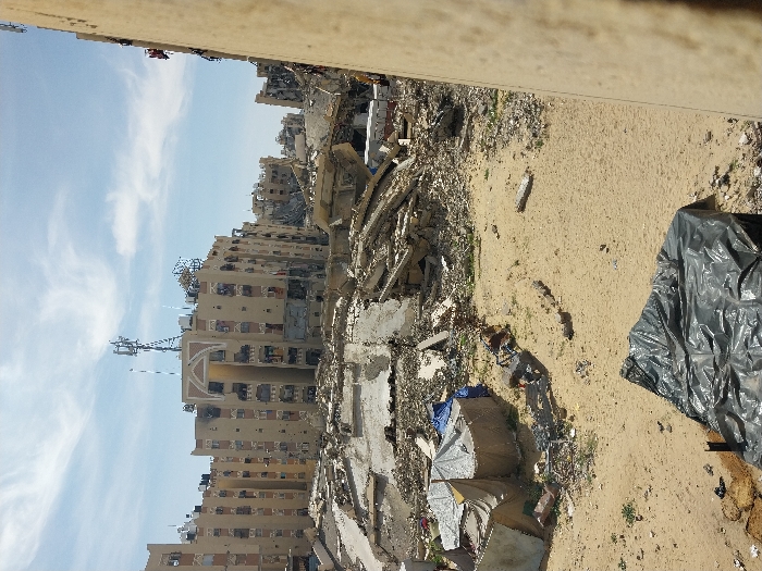 An outdoor, wide-angle image depicts a scene of extensive structural damage in the city of Khan Yunis, Palestinian Territory, under a bright, partly cloudy sky. The foreground features sandy ground with sparse vegetation. In the lower left, a dark grey or black tarp covers an unseen object. To the left-center, three makeshift shelters, constructed from light-colored fabric and blue tarpaulin, stand on the sandy ground amidst scattered debris.
The mid-ground is dominated by massive piles of concrete rubble, twisted rebar, and fragments of collapsed buildings, indicating widespread destruction. In the background, several multi-story buildings, appearing residential or commercial, are heavily damaged but partially standing. One prominent building in the center-left background features an arched design element on its upper facade and a communication tower with multiple antennas on its roof; this building exhibits visible structural damage and darkened areas. Other partially visible buildings further back also show signs of significant destruction. The right edge of the frame is bordered by a light-colored, vertical wall or structure. No individuals are visible in the scene.