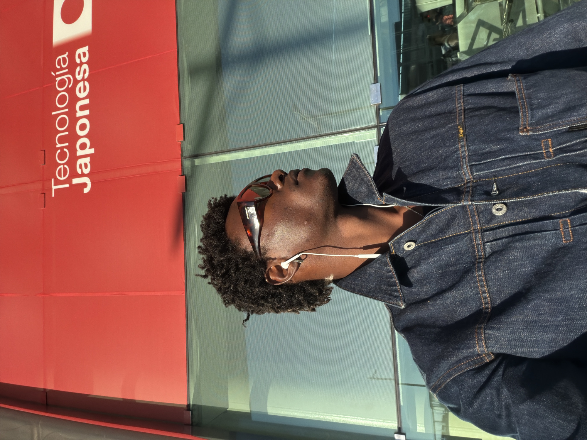 A medium, eye-level shot depicts a man wearing a dark denim jacket and sunglasses standing next to a building. He is wearing white earbuds and looking upwards. The building has a red panel with the text "Tecnologia Japonesa" and a stylized Japanese flag on it. The building also has glass panels that reflect the surroundings. The photograph was taken in the city of El Prat de Llobregat, Spain.
