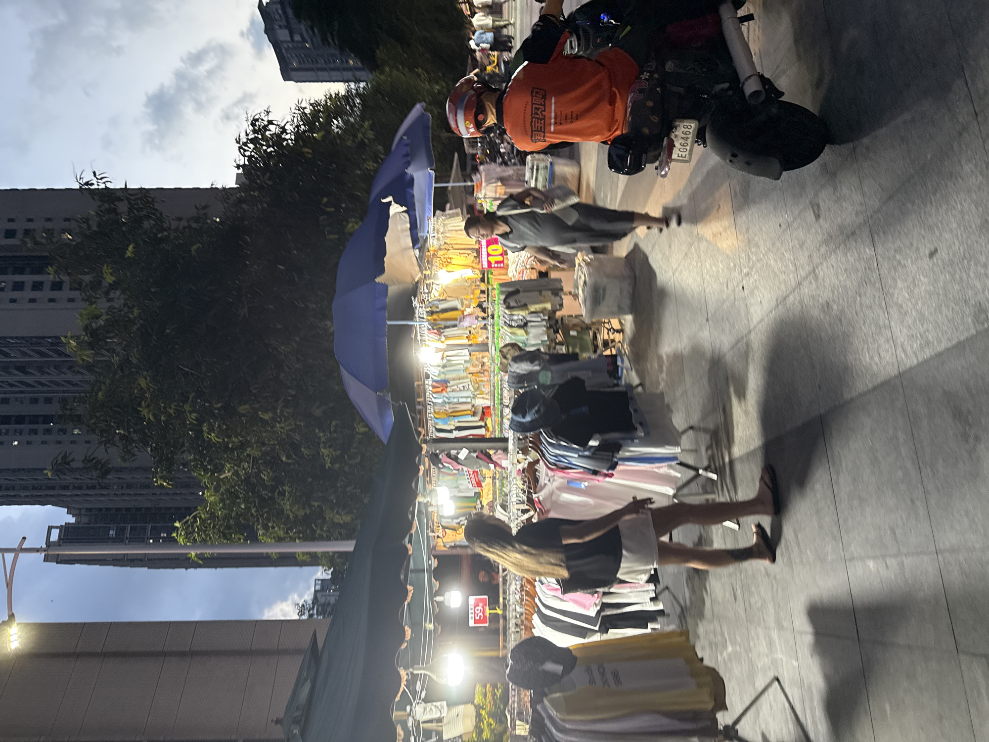 The image depicts a street scene at dusk in Haizhu District, China. A woman with long dark hair is browsing clothing racks at an outdoor market. She is wearing a black t-shirt and white shorts. In the background, a woman in a grey dress walks past a stall displaying various garments. To the right, a person on a black scooter wears an orange jacket and a red helmet. The scooter has a license plate reading "EG6468". Overhead, a blue umbrella covers some of the stalls. The ground is paved with grey tiles. In the distance, tall buildings rise against a twilight sky with scattered clouds. A street lamp illuminates the area on the left.