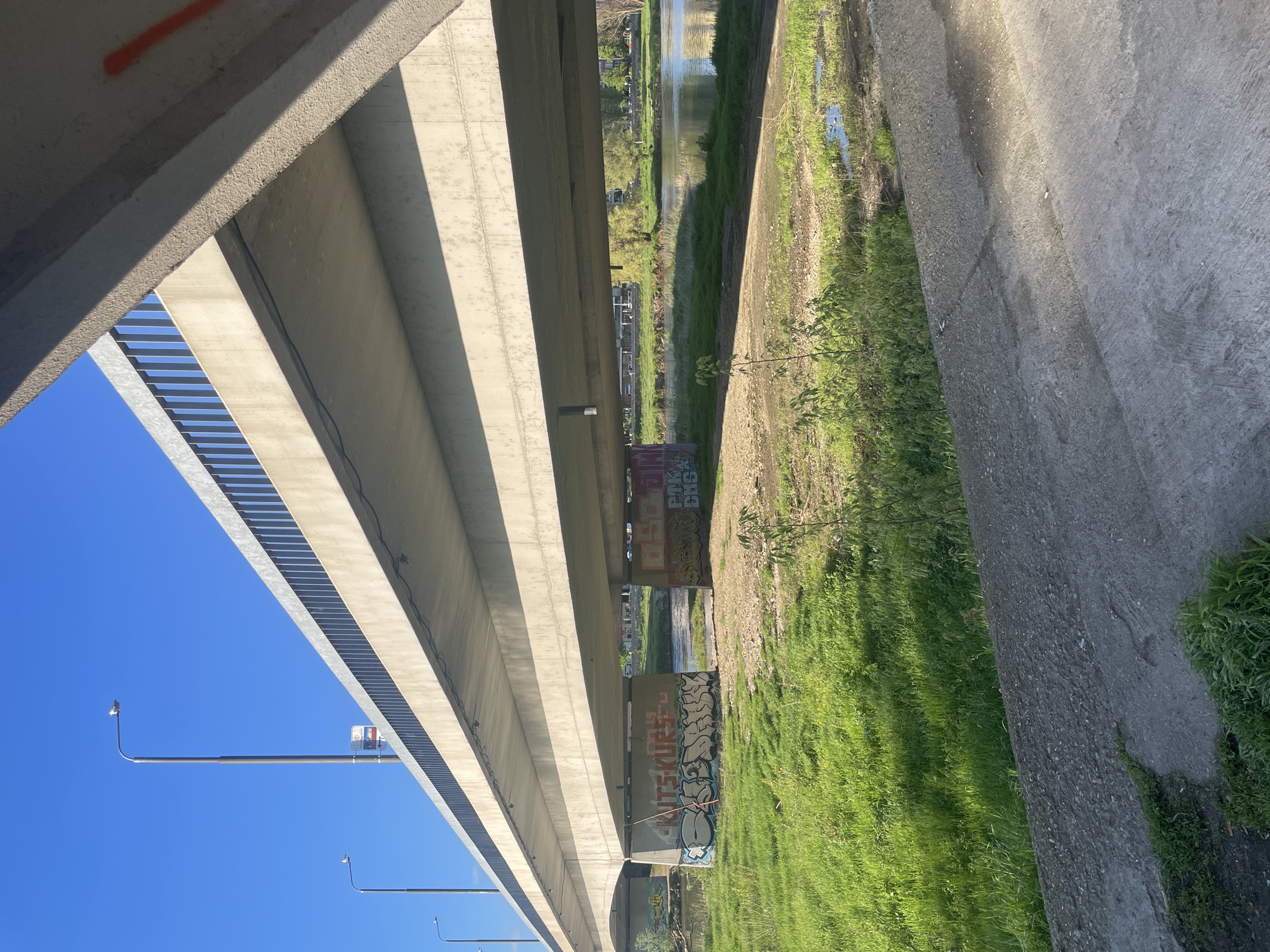 The image, taken from a low angle, depicts the underside of a large concrete bridge spanning across a waterway and adjacent land in Plovdiv, Bulgaria. The massive concrete structure of the bridge, with its prominent girders and support columns, dominates the view. Sunlight creates sharp, elongated shadows across the concrete surfaces, emphasizing the bridge's geometric lines and textures.
Beneath the bridge, a body of water, likely a river or canal, flows through an open area. Its surface reflects the sky and surrounding greenery. On both sides of the water, the ground is covered in vibrant green grass, interspersed with patches of bare earth and some small bushes or saplings.
A clear, bright blue sky is visible through the openings in the bridge's framework. Several streetlights with long, slender arms are mounted on the bridge structure, extending upwards into the open sky. A metal safety railing can also be seen along the side of the bridge.
Notable details include graffiti spray-painted on the concrete support columns under the bridge. Visible text includes "POK CREW" and "KITSKORE," alongside other indistinct tags and designs. A small orange marking is also present on an upper concrete beam. The scene is set on a clear, sunny day, with no people or explicit activity depicted, contrasting the urban infrastructure with the natural environment.