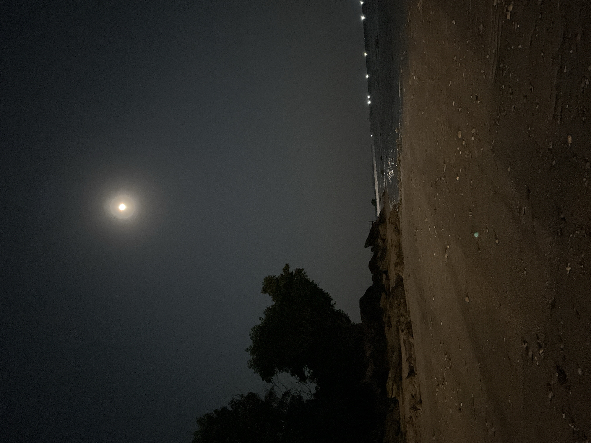 This is a night-time, landscape-oriented photograph taken on a beach in Legonipah, Indonesia. The primary subjects are the moon, the ocean, the beach, and some vegetation.
The moon is a prominent, luminous orb in the dark sky, casting a soft glow. To the right of the moon, in the distance, is a line of lights, likely from a town or boats on the water. The ocean stretches across the upper portion of the image, with some gentle waves visible near the shore. The beach occupies the right side of the frame, appearing sandy with scattered stones and debris. A dark silhouette of trees and other vegetation is visible along the edge of the beach, contrasting with the lighter sand and sky.
The overall mood of the image is serene and tranquil, characteristic of a quiet night by the sea. The time of day is clearly night due to the presence of the moon and the darkness. The location cues, such as the beach and the ocean, along with the provided context of Legonipah, Indonesia, help to establish the setting. There is no visible text in the image.