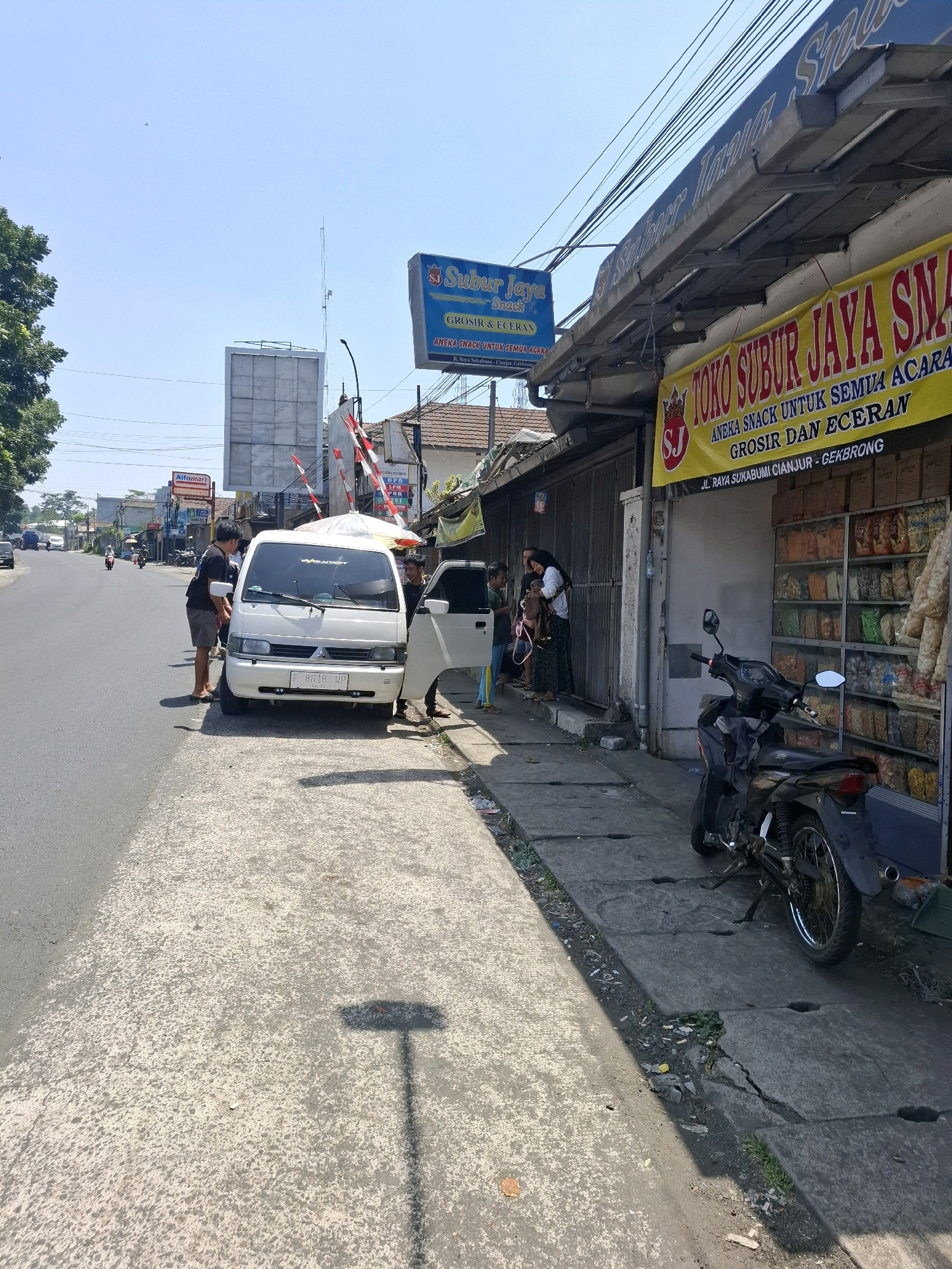 The image depicts a street scene in Cikahuripan, Indonesia, on a clear, sunny day. In the foreground, a gray asphalt road is visible on the left, with a concrete sidewalk to its right. Several people are present. Two men stand near a white minivan parked on the side of the road. One man, wearing a dark t-shirt and shorts, is leaning towards the minivan's driver's side. Another man, also in dark clothing, stands by the open passenger door of the minivan. Further down the sidewalk, a group of people, including women and children, are gathered near a storefront.
The storefront is a shop, identified by a yellow banner with red text that reads "TOKO SUBUR JAYA SNA". Below this, smaller text states "ANEKA SNACK UNTUK SEMUA ACARA GROSIR DAN ECERAN JL. RAYA SUKABUMI CIANJUR - CEKBRONG". The shop has a glass display showcasing numerous bags of snacks. To the right of the shop, a black motorcycle is parked on the sidewalk.
In the background, the street extends into the distance, lined with buildings and trees. A large billboard or structure is visible, along with a radio tower. Traffic signals and streetlights are also present. The overall impression is a bustling local street with commercial activity.