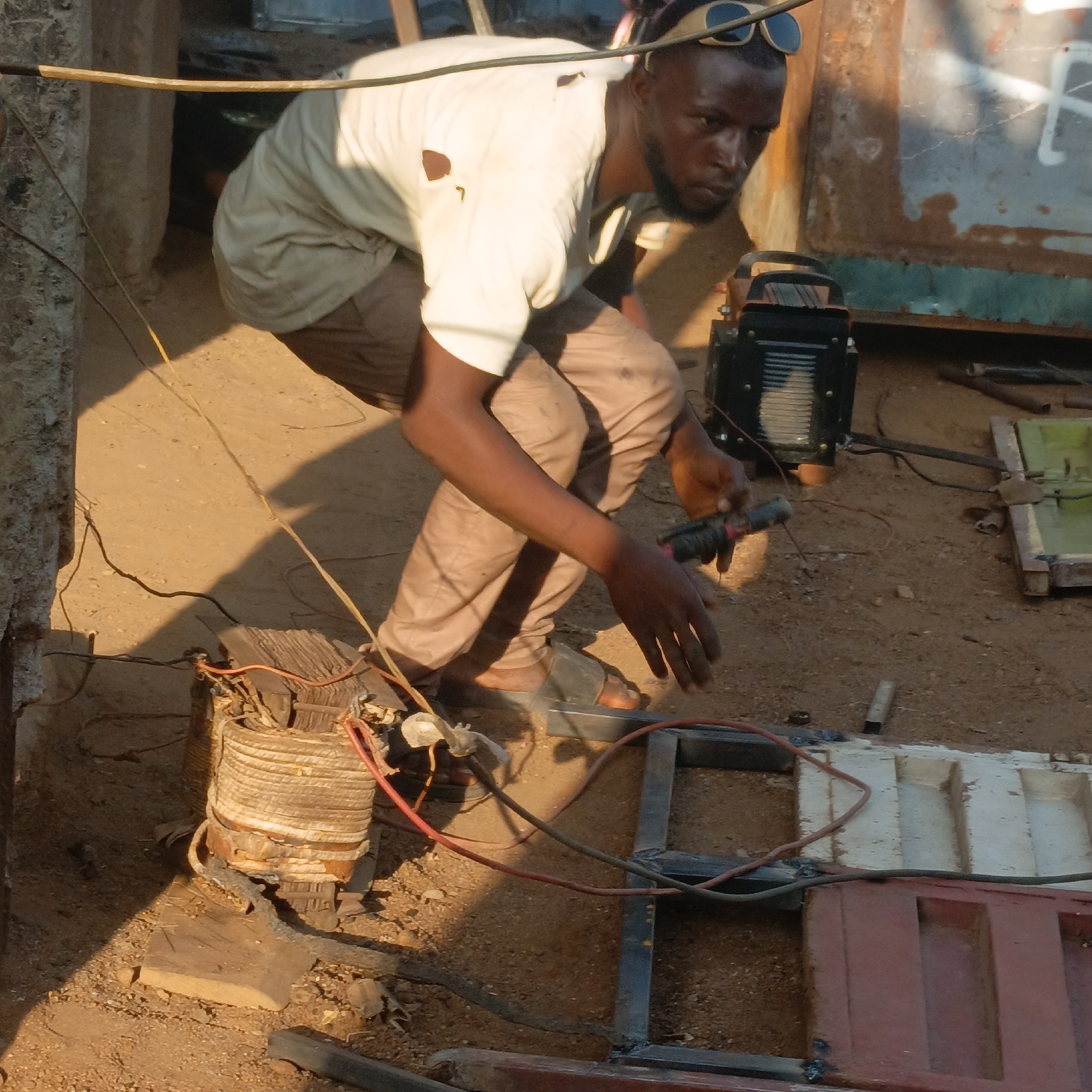 In Akwanga, Nigeria, an adult male is squatting outdoors, holding a welding tool near a partially constructed metal frame. He wears a light-colored shirt with a tear, brown pants, and sandals. Sunglasses are perched on his head. A welding machine sits to his right, connected to the transformer by wires. The ground is dusty and there is a concrete wall fragment nearby. A metal object lies on the metal frame that is being constructed.