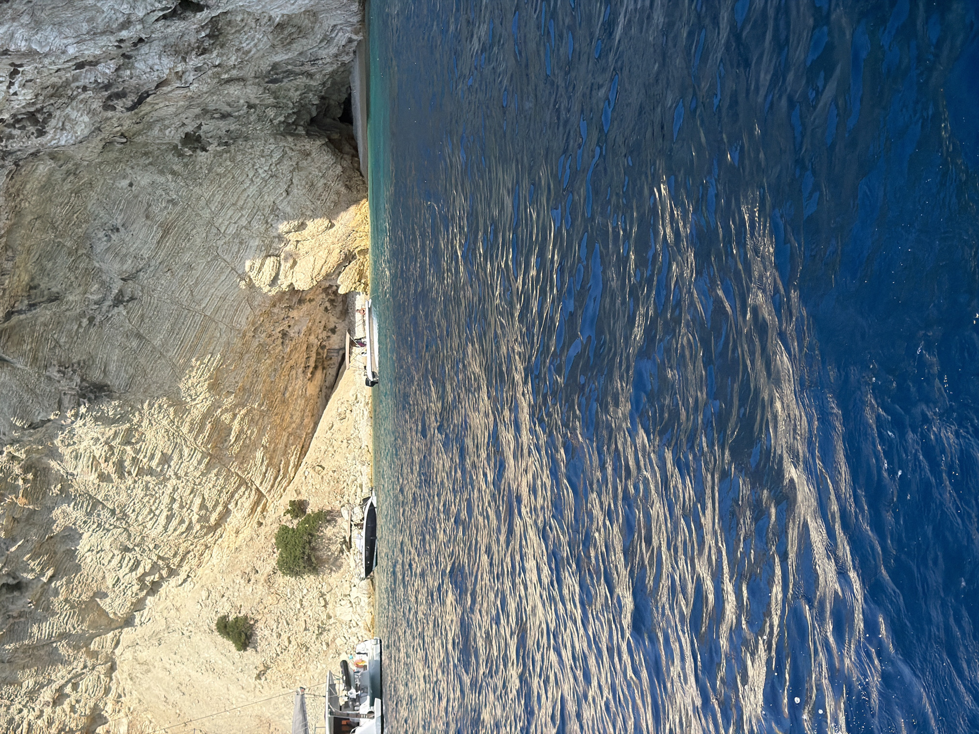 The image shows a body of water in front of a rocky cliff face. There are several boats visible in the water, and some people are on one of the boats. The cliff face has a cave or opening in it. The water is a deep blue color and appears somewhat choppy. The cliff is a light brown or tan color and appears to be made of layered rock. There are some small green bushes growing on the cliff face. The scene is lit by bright sunlight, casting shadows on the cliff. This image appears to be taken from the sea looking towards the shore.