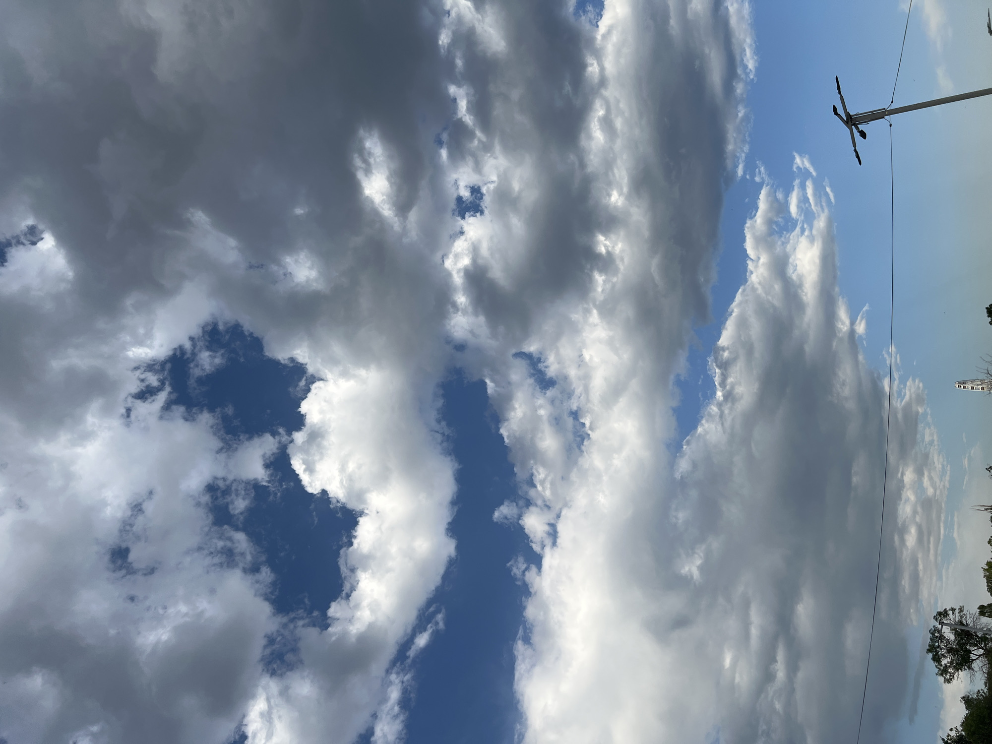 This is an image of a cloudy sky over Yerevan, Armenia. The sky is filled with both white and dark grey clouds, with patches of blue sky visible in between. In the lower right corner of the image, a utility pole with multiple lights is visible. In the distance, at the horizon, a Ferris wheel can be seen. The lower portion of the image shows the tops of some trees and a line of what appears to be power lines stretching across the frame. The lighting suggests it may be late afternoon or early evening. There are no people visible in the image.