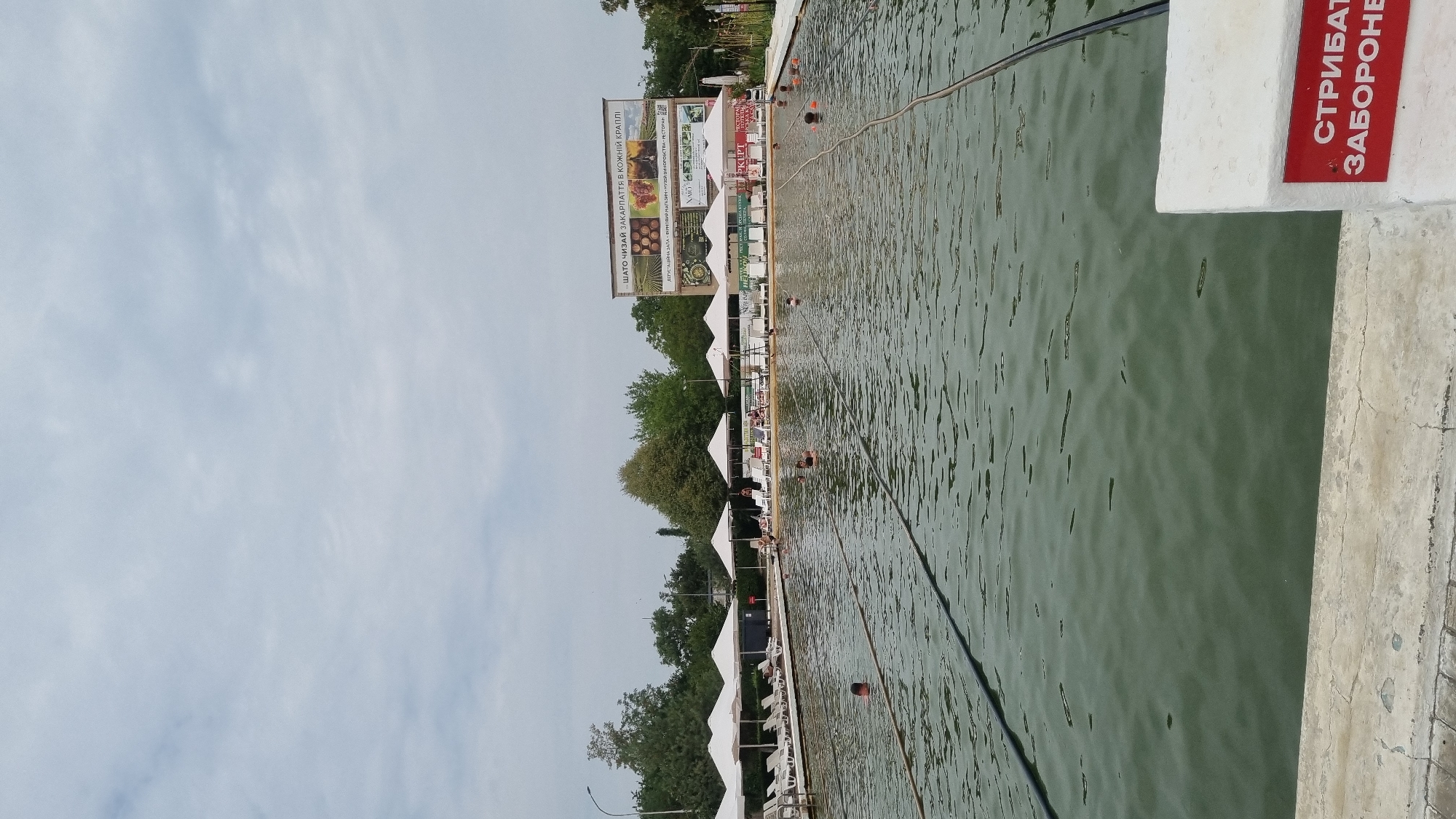 The image shows a large outdoor swimming pool with murky green water, possibly a thermal pool, located in Berehove, Ukraine. Several people are swimming or wading in the pool. Along the left side of the pool, there is a row of white lounge chairs and umbrellas. In the background, there are trees and a large billboard with text and images. On the right side of the foreground, a white pillar with a red sign displaying white text in Cyrillic script is visible. The sky is overcast.