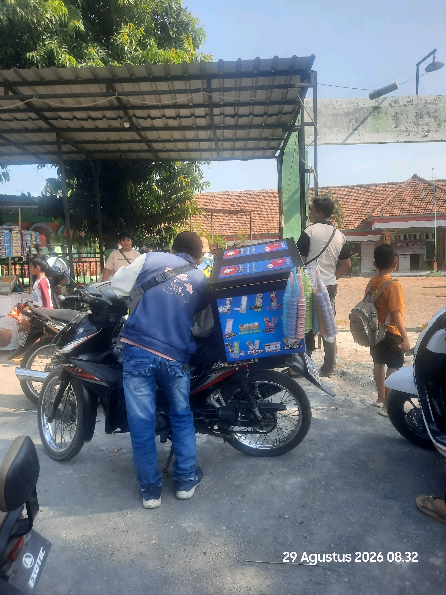 The image shows a sunny outdoor scene with several people and motorcycles in Sukokerto, Indonesia. In the foreground, a person in a blue jacket and jeans stands next to a motorcycle equipped with a large, dark blue cooler box. The cooler box is adorned with colorful illustrations and text, suggesting it is used for selling frozen treats or drinks. Several colorful ice cream cups are attached to the side of the cooler.
To the left, another motorcycle is parked, and a young person in a white and red shirt is visible standing behind it. Further back, two young men are standing near the cooler box. One is wearing a light-colored t-shirt and has a strap across his chest, while the other is wearing a dark t-shirt. A young boy in an orange t-shirt and backpack stands to the right of the cooler.
In the background, there is a covered structure with a tiled roof and a metal frame, possibly a market stall or a small building. Beyond that, there are buildings with red tiled roofs. The ground is a paved surface, appearing to be asphalt or concrete, with some scattered debris. The overall atmosphere suggests a casual, everyday scene in a public area, likely a street or marketplace. The timestamp in the lower right corner indicates the image was taken on August 29, 2026, at 08:32.