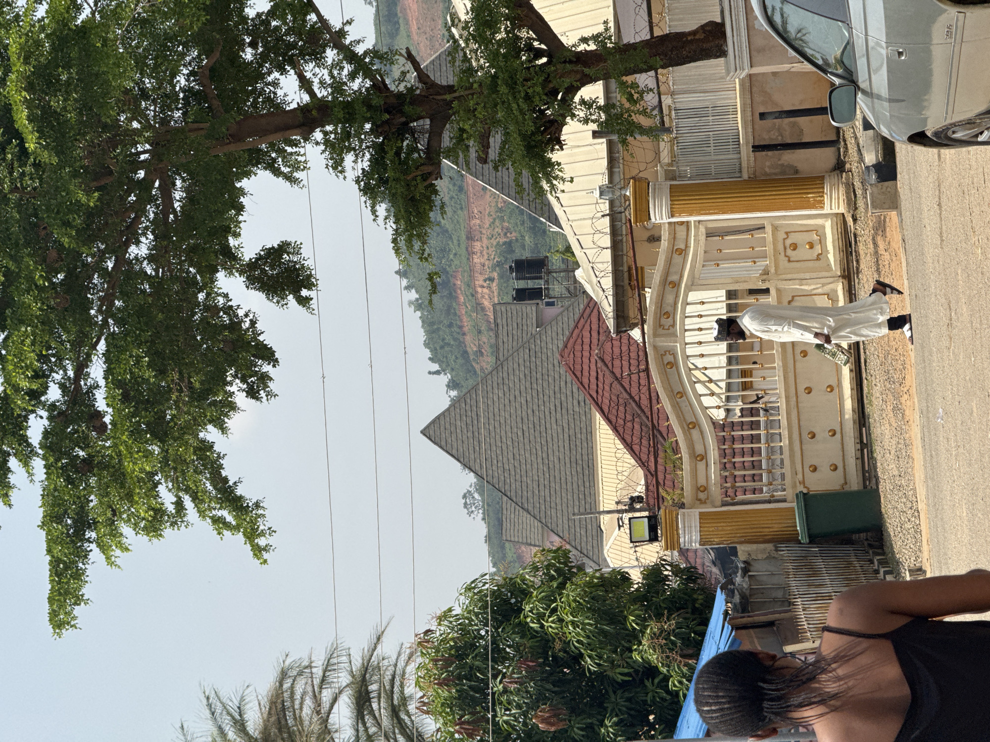 The image depicts an outdoor scene with a man in traditional Nigerian attire walking past a white and gold gate. He is wearing a white robe and a cap, and he is holding a small green object in his right hand. Behind him, a silver car is parked on the side of the road. The gate is ornate, with decorative gold pillars and white panels. A large tree with green leaves shades part of the scene. In the background, there are several houses with red and grey roofs, and a hilly landscape covered in greenery and some brown earth. The sky is clear and bright, suggesting it is daytime, likely late morning or early afternoon, with fair weather. Power lines are visible crossing the scene. The overall impression is one of a residential area in Abuja, Nigeria, on a clear day.