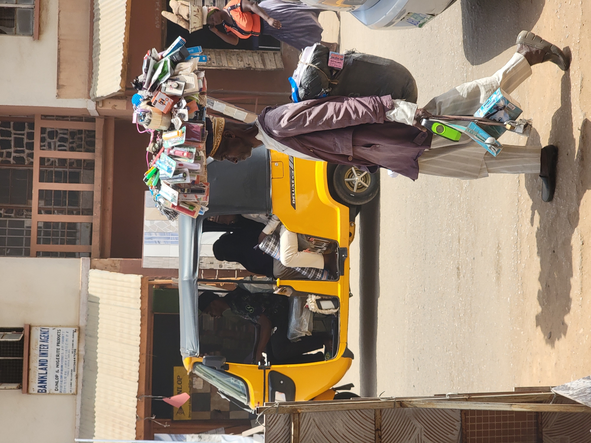 The image captures a bustling street scene in Maiburiji, Nigeria, featuring a man carrying a large assortment of goods balanced on his head. He is walking alongside a yellow "Deluxe" tricycle taxi, commonly used for local transportation. The man is dressed in striped pants, a dark shirt, a brown jacket and a head piece. He's carrying tools on the side. Passengers can be seen inside the taxi. In the background, there are buildings with windows, and a sign for "BANKLAND INTER AGENCY DUNLOP & NIGERITE PRODUCTS." A person wearing an orange life vest also appears in the background. The scene unfolds in daylight on a paved road.