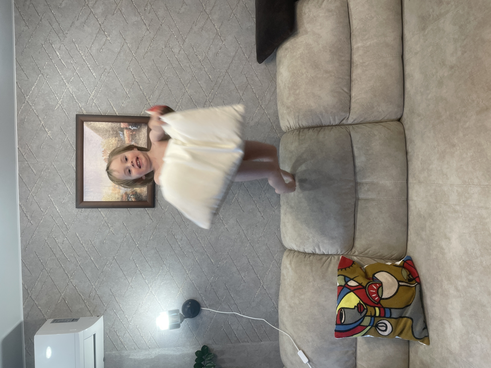 A young child, possibly a toddler, is captured mid-jump in an indoor setting, holding a large white pillow. The child has blonde hair and is looking towards the camera with a playful expression. They are wearing only a diaper. The background features a textured wallpaper with a subtle geometric pattern. On the wall, a framed picture hangs, and to the left, an air conditioning unit and a wall-mounted light fixture are visible. In the foreground, a beige sectional sofa occupies the lower portion of the frame. On the left side of the sofa, a decorative pillow with a colorful abstract design is placed. The lighting suggests an indoor environment, possibly a living room. No specific city or location details are discernible from the image itself.
