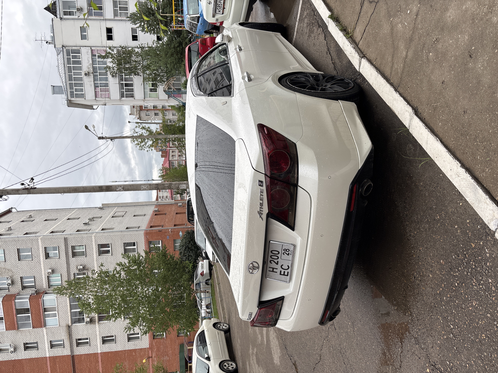 The image captures a white Toyota Crown Athlete S parked on a wet asphalt surface in an urban setting. The car is positioned at an angle, with its rear visible to the viewer. The license plate reads "H 200 EC 28 RUS". The car is detailed with a chrome trim around the windows and black multi-spoke rims. The surroundings include apartment buildings, trees, and a cloudy sky. The presence of a playground structure and other parked vehicles suggests a residential area. The overall scene indicates a cloudy or rainy day, given the wetness of the ground and the droplets visible on the car's surface. The architecture of the buildings suggests a location in Blagoveshchensk, Russia, characterized by typical Soviet-era apartment blocks.