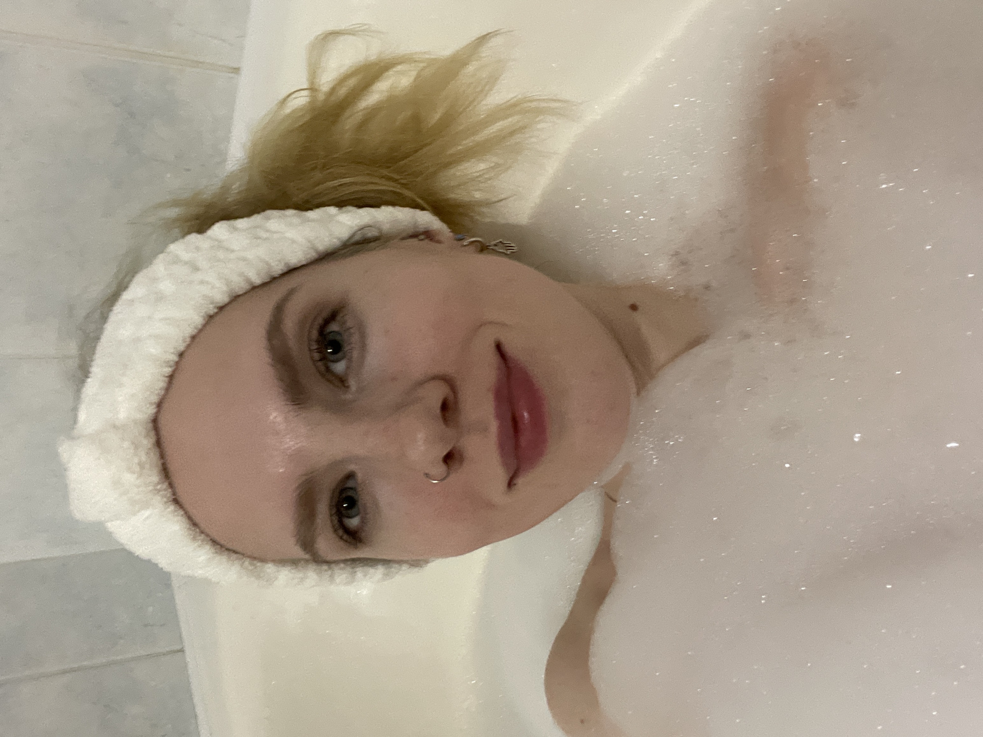 A person is in a bathtub filled with bubbles. The person has fair skin, blonde hair pulled up in a bun, and is wearing a white, terrycloth headband. The person's face is turned towards the camera with a slight smile. A small, silver nose ring is visible in the left nostril. A tiled wall is partially visible in the background on the left. The person is lying in a tub filled with white soap bubbles. There is no indication of location in the city of Izhevsk, Russia.