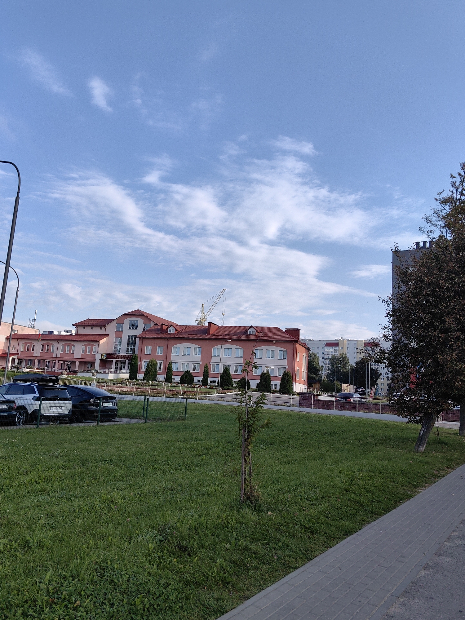 A wide shot captures a pink, multi-story school building with a red-tiled roof under a blue sky with wispy white clouds. A large crane is visible behind the school building, and several cars are parked in front of a low fence. The foreground is dominated by a green lawn, with a gray paved walkway running along the right side of the image. A street lamp is visible on the left side of the frame. The school has Cyrillic text above its entrance. The scene is located in Fanipal, Belarus.