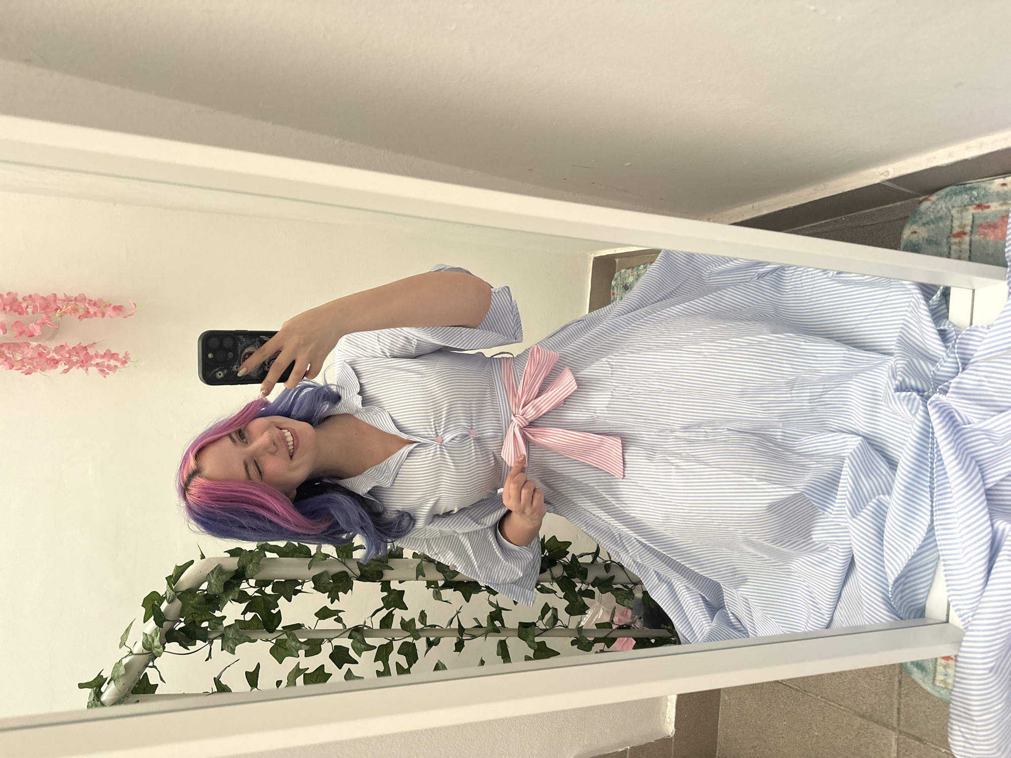 A young woman is captured taking a mirror selfie in an indoor setting, located in the City of Zagreb, Croatia. She is smiling broadly and looking into the smartphone she holds up with her right hand to capture the reflection. Her hair features distinct streaks of pink and blue/purple. She is dressed in a light blue and white striped shirt dress, which has a prominent pink and white striped bow tied around the waist.
The selfie reveals white walls in the background. To the left, a cluster of pink artificial flowers, resembling cherry blossoms, is visible. Below the woman, an artificial green ivy garland is draped over a white railing or decorative frame. The bottom edge of the white mirror frame is visible across the lower part of the image. A section of a patterned rug or carpet, featuring blue, white, and reddish tones, can be seen on the right side, just outside the mirror's main reflection but still in the camera's view. The smartphone she is holding is black, equipped with multiple camera lenses, and encased in a decorative cover that appears to have a dark, indistinct design. The lighting suggests it is daytime, and the woman's expression conveys happiness. No visible text is present in the image.