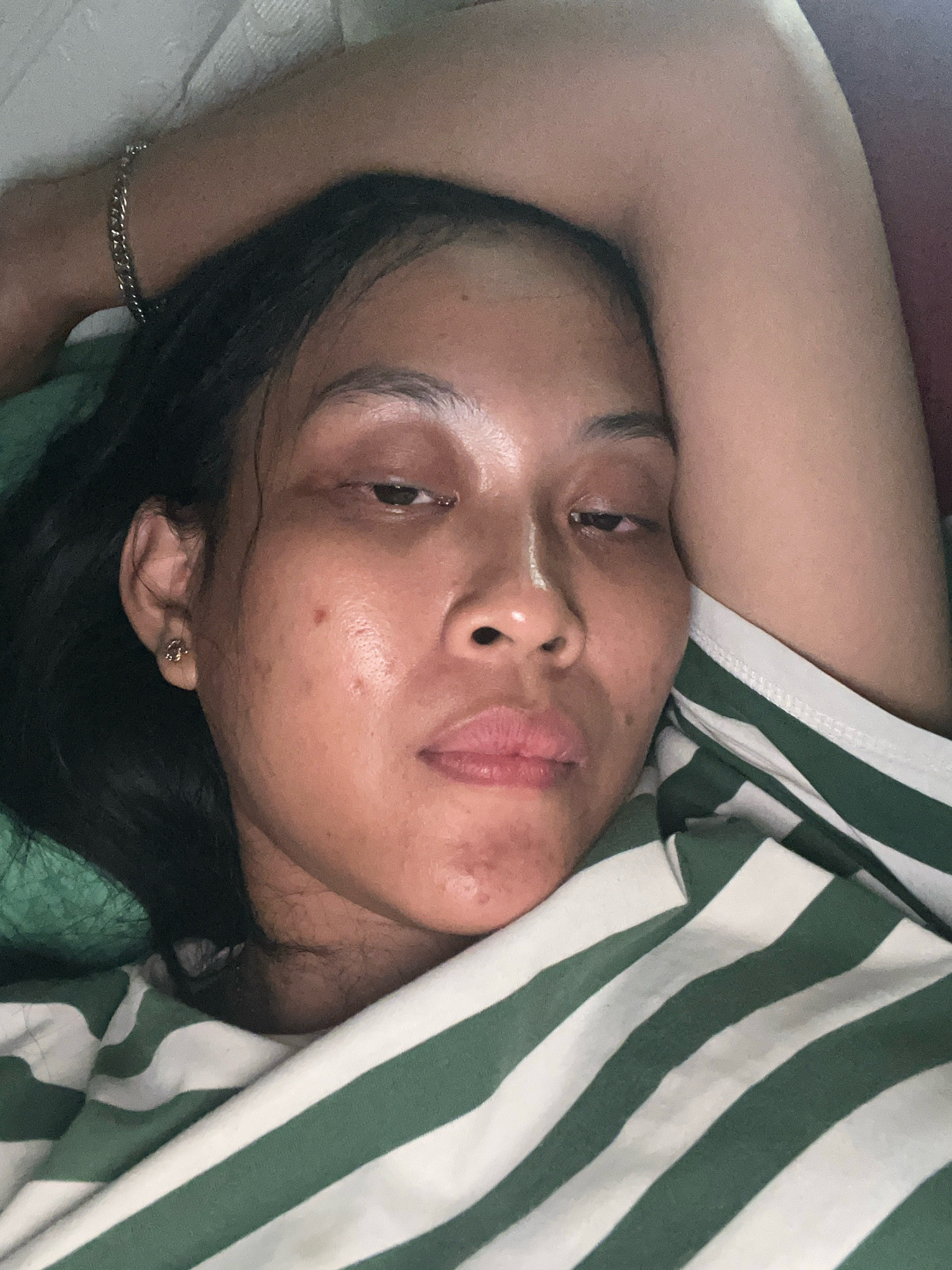 A close-up, overhead shot captures a young woman lying down, her face turned towards the camera, with her left arm bent behind her head. She is wearing a green and white striped shirt. Her eyes are partially closed, and her expression is relaxed. A silver bracelet adorns her left wrist. The background is out of focus, but suggests an indoor setting. The image was taken in Kecamatan Pati, Indonesia.