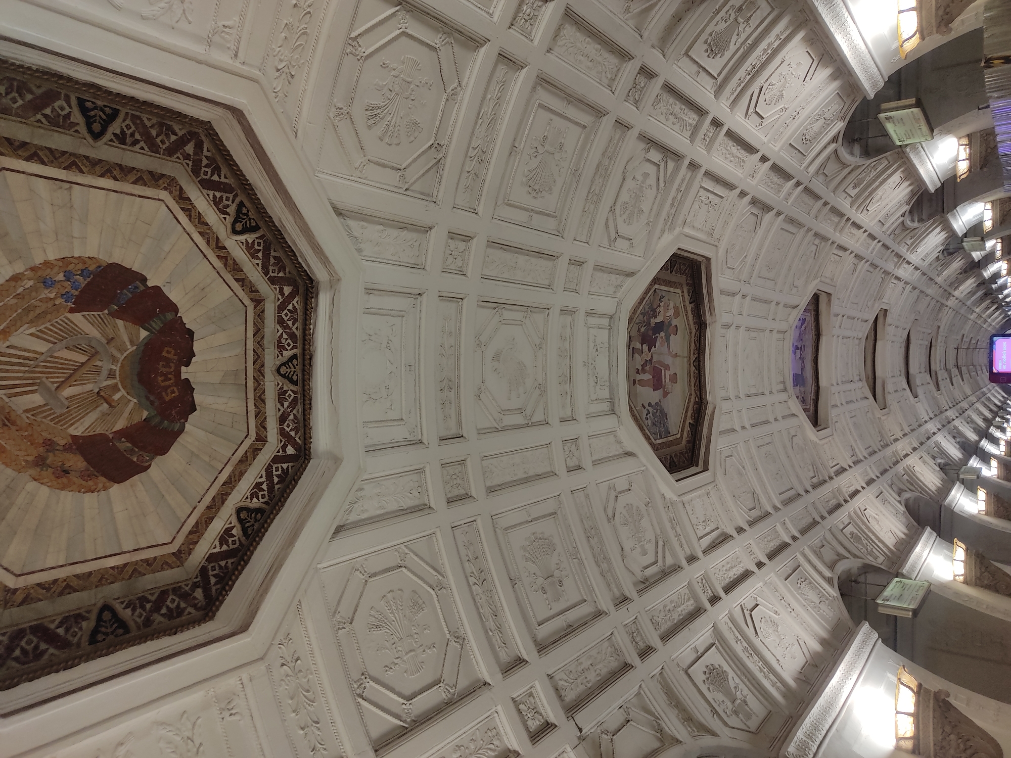 The image shows a highly ornate, arched ceiling in the Kiyevskaya station on the Arbatsko-Pokrovskaya Line of the Moscow Metro in Moscow, Russia. The ceiling is a series of recessed, octagonal sections with intricate relief patterns and occasional mosaics or paintings. Lighting fixtures are visible on the side walls. The overall impression is one of elaborate decoration and architectural grandeur.