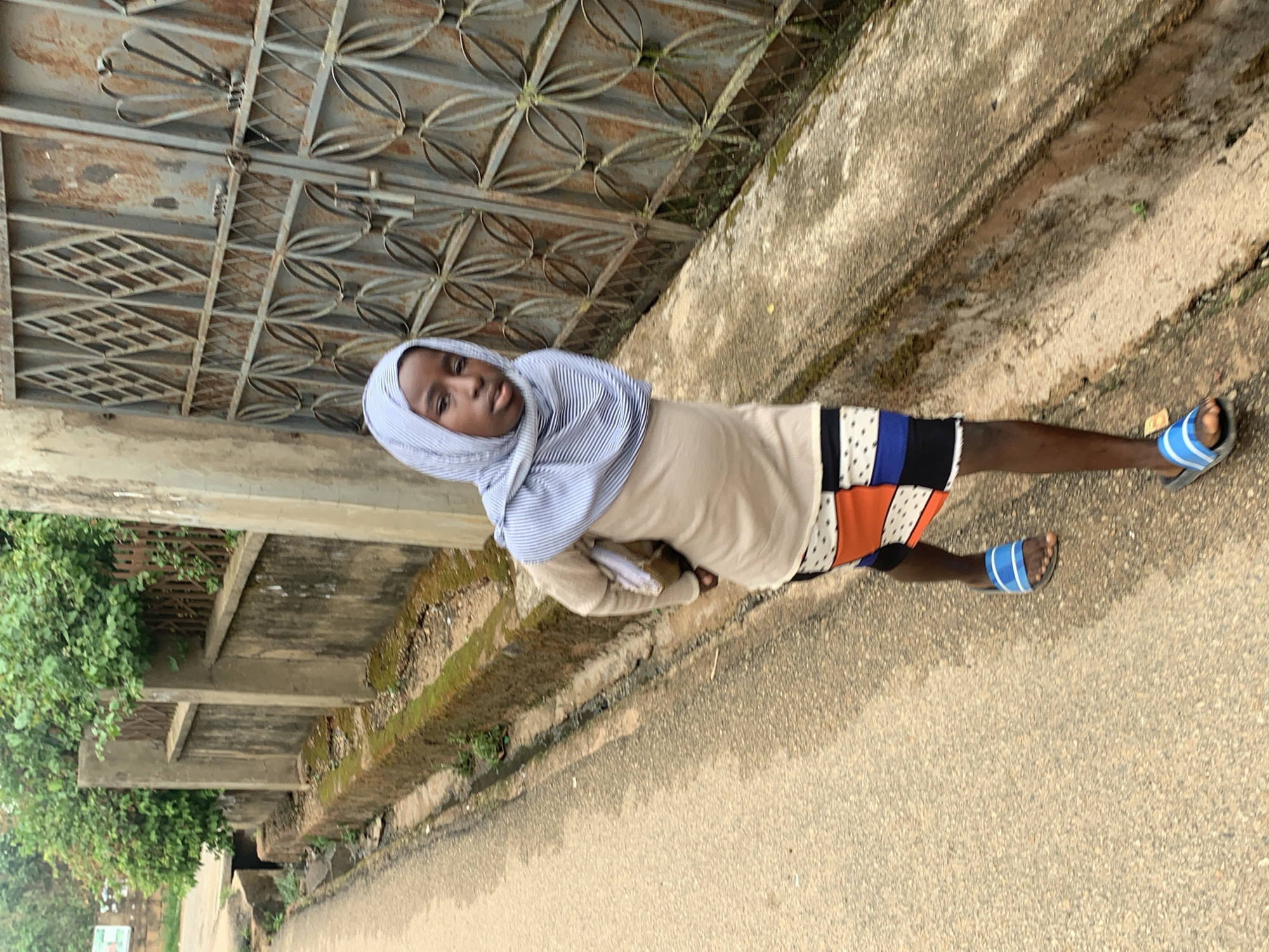 A person is standing on a sidewalk in Jos, Nigeria. The person is wearing a headscarf, a beige shirt, and a skirt with multiple colors and patterns. They are also wearing blue sandals. The sidewalk is next to a road. There is a wall and a metal gate to the left of the person. Greenery can be seen behind the wall. The scene is well-lit with natural light.