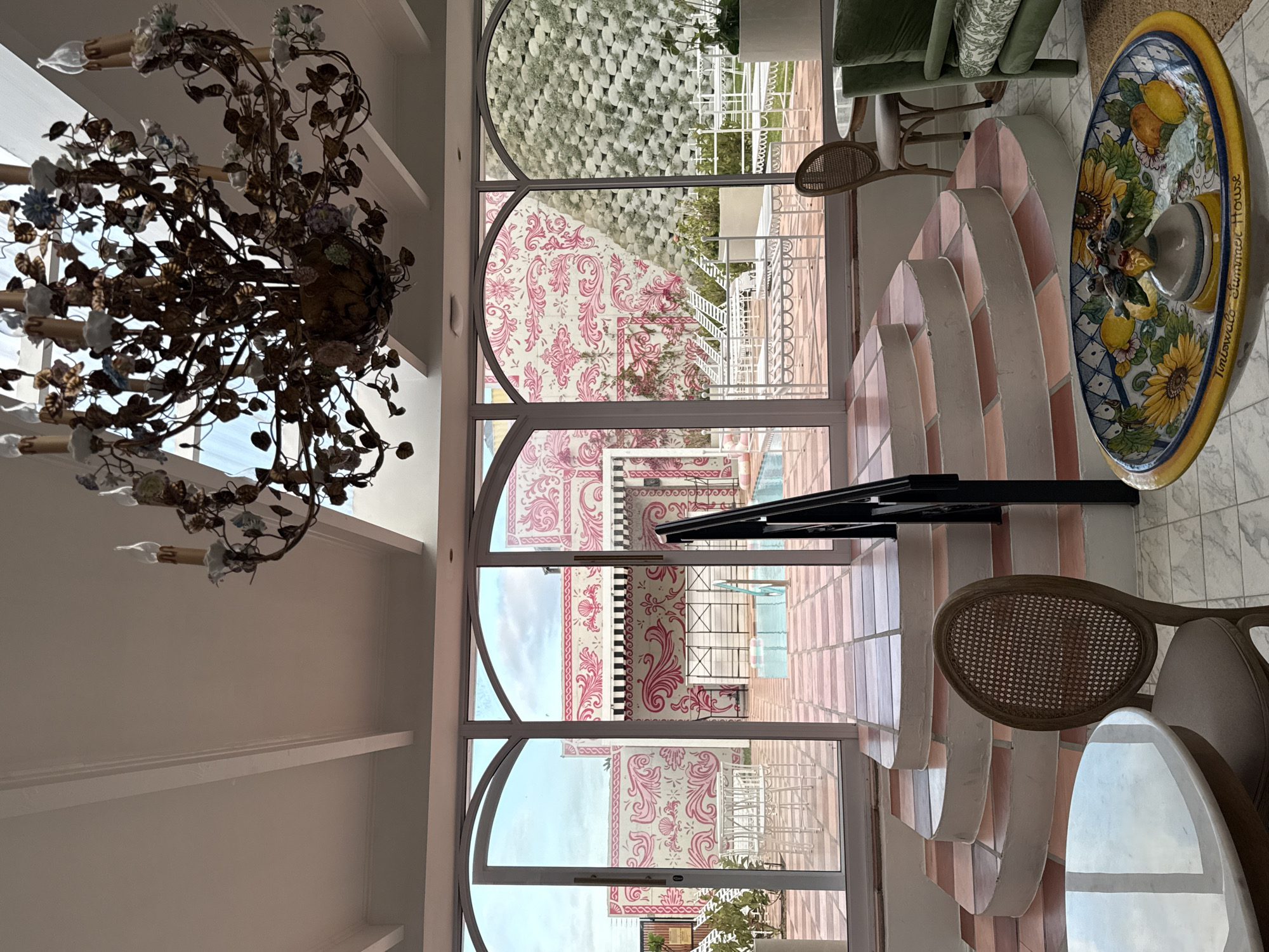 The image shows an interior view of a building with a prominent chandelier in the foreground. Through a large arched window or doorway, there is a view of the outside with patterned pink and white building exterior, a swimming pool, and what appears to be a tiled patio. There are chairs and a round decorative plate or platter on a table inside. The exterior's architectural style suggests a whimsical or decorative design, and the patterned walls are a striking feature.
The location is in the city of Cape Town, South Africa.