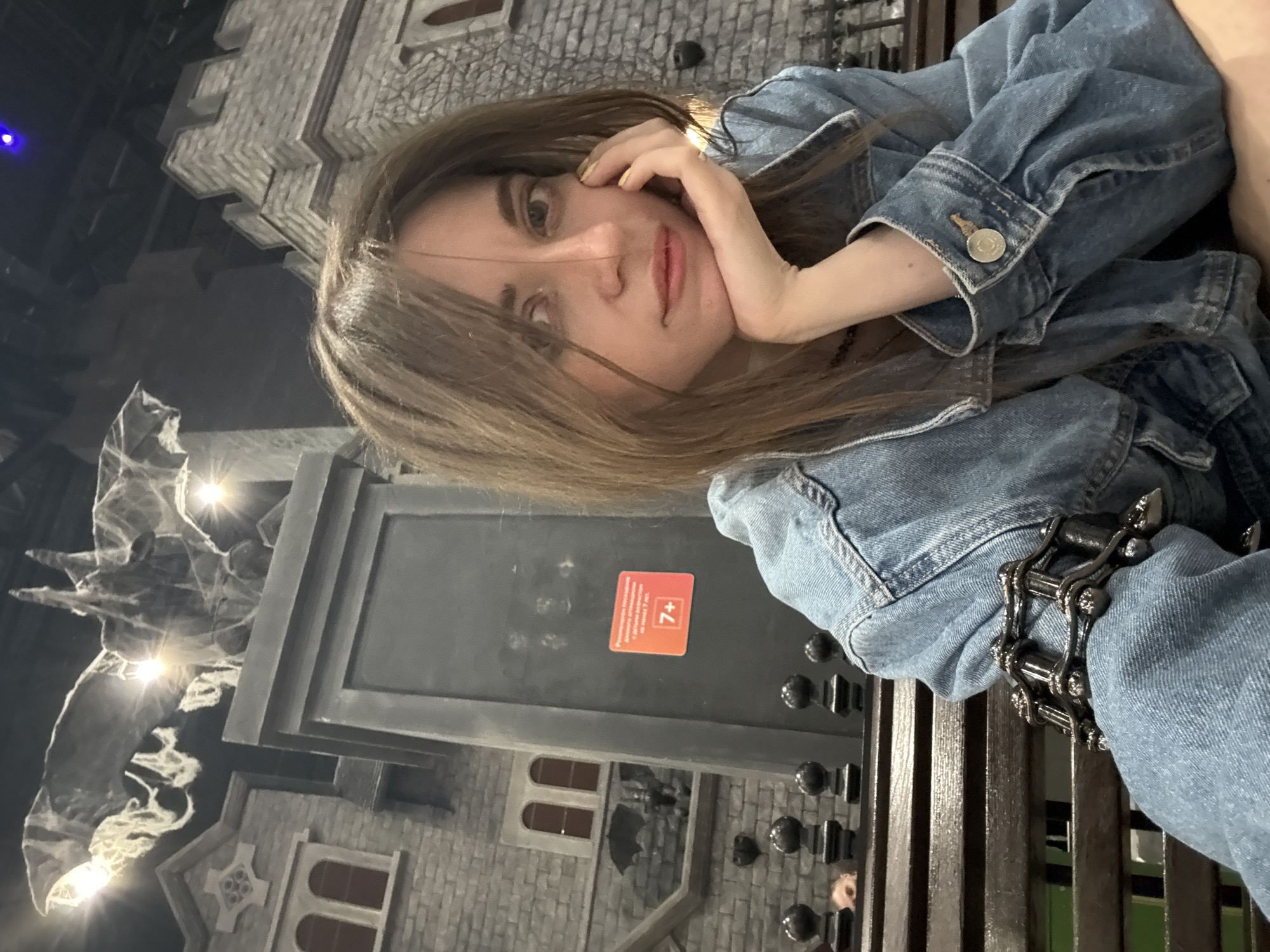 A woman is seated, wearing a denim jacket and a black bracelet. She rests her head on her hand and looks at the camera. In the background is a castle-themed area, likely for children's entertainment, with a sign indicating it's appropriate for ages 7+. The location is Moscow, Russia.
