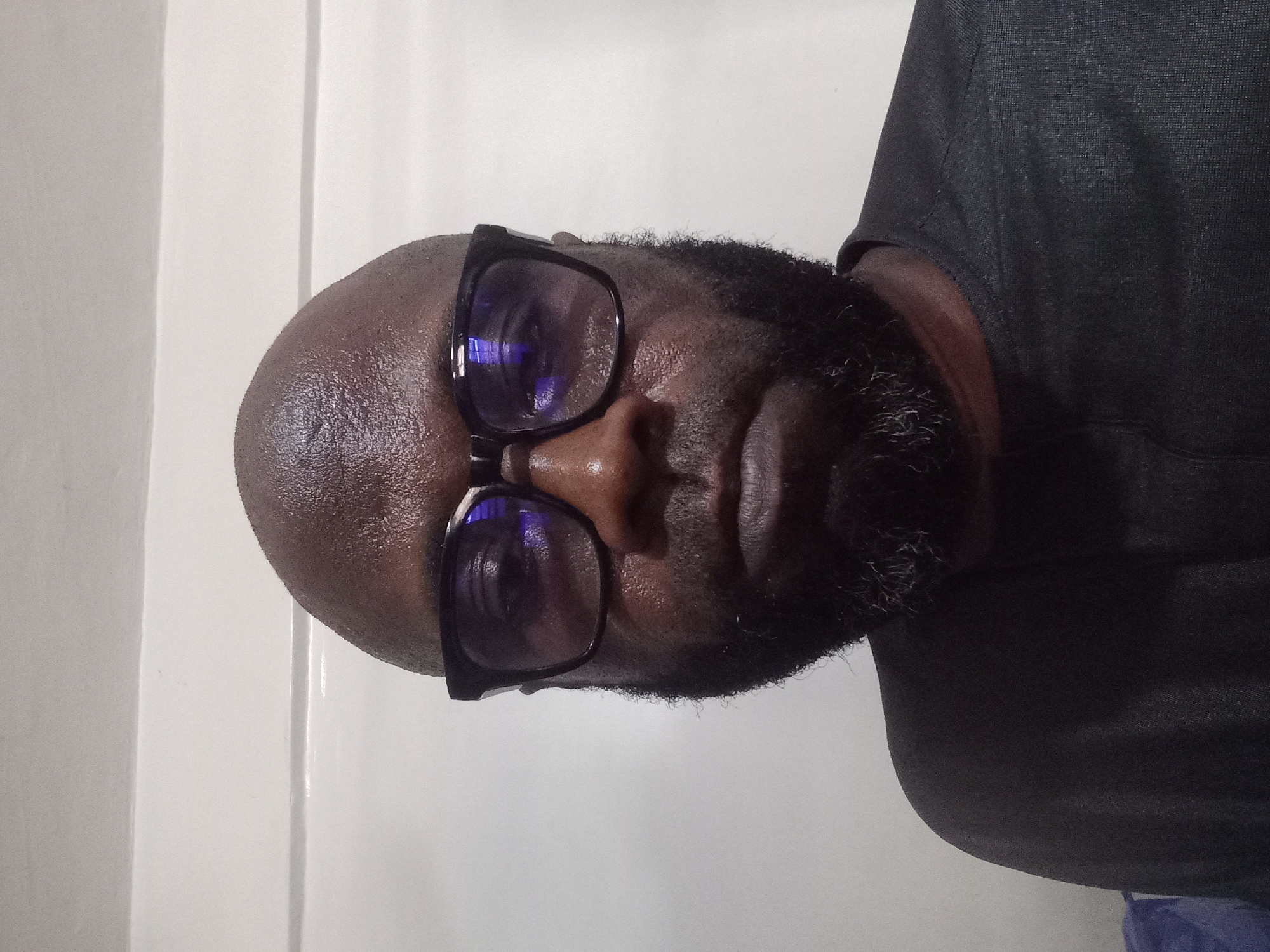 An image shows a man wearing glasses with a black frame. He has a bald head and a beard. He wears a black t-shirt. The background is a white wall. The image does not show any information about the city of Dadi Riba, Nigeria.