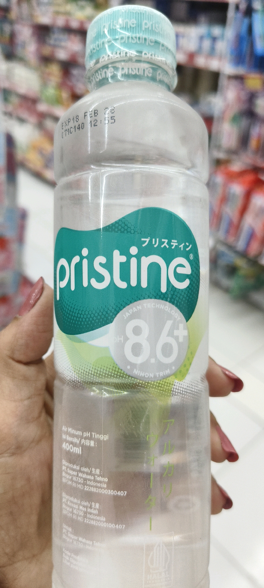 A person's hand with pink nail polish is holding a clear plastic bottle of Pristine brand alkaline water. The bottle has a teal label with white text and graphics. The label prominently displays "pristine" in large white letters. Above this, in smaller Japanese text, it says "プリスティン". There is a circular graphic with "JAPAN TECHNOLOGY" and "8.6+" within it, and "NIHON TRIM" below. Further down the bottle, in smaller white text, it states "Air Minum pH Tinggi" (High pH Drinking Water), "Isi Bersih/ 内容量:" (Net Content:), and "400ml". Below this are production details, including "Diproduksi oleh/ 生産:" (Produced by/Manufactured:), "PT. Super Wahana Tehno", "Bogor 16730 - Indonesia", and "BPOM RI MD 222882000300407". Another production entry for "PT. Kreasi Mas Indah" is also visible. The background is blurred and appears to be a store aisle with shelves stocked with various products. The overall impression is a product shot of the water bottle in a retail setting. The image does not provide information about the city of Waisai, Indonesia, or any specific location within it.