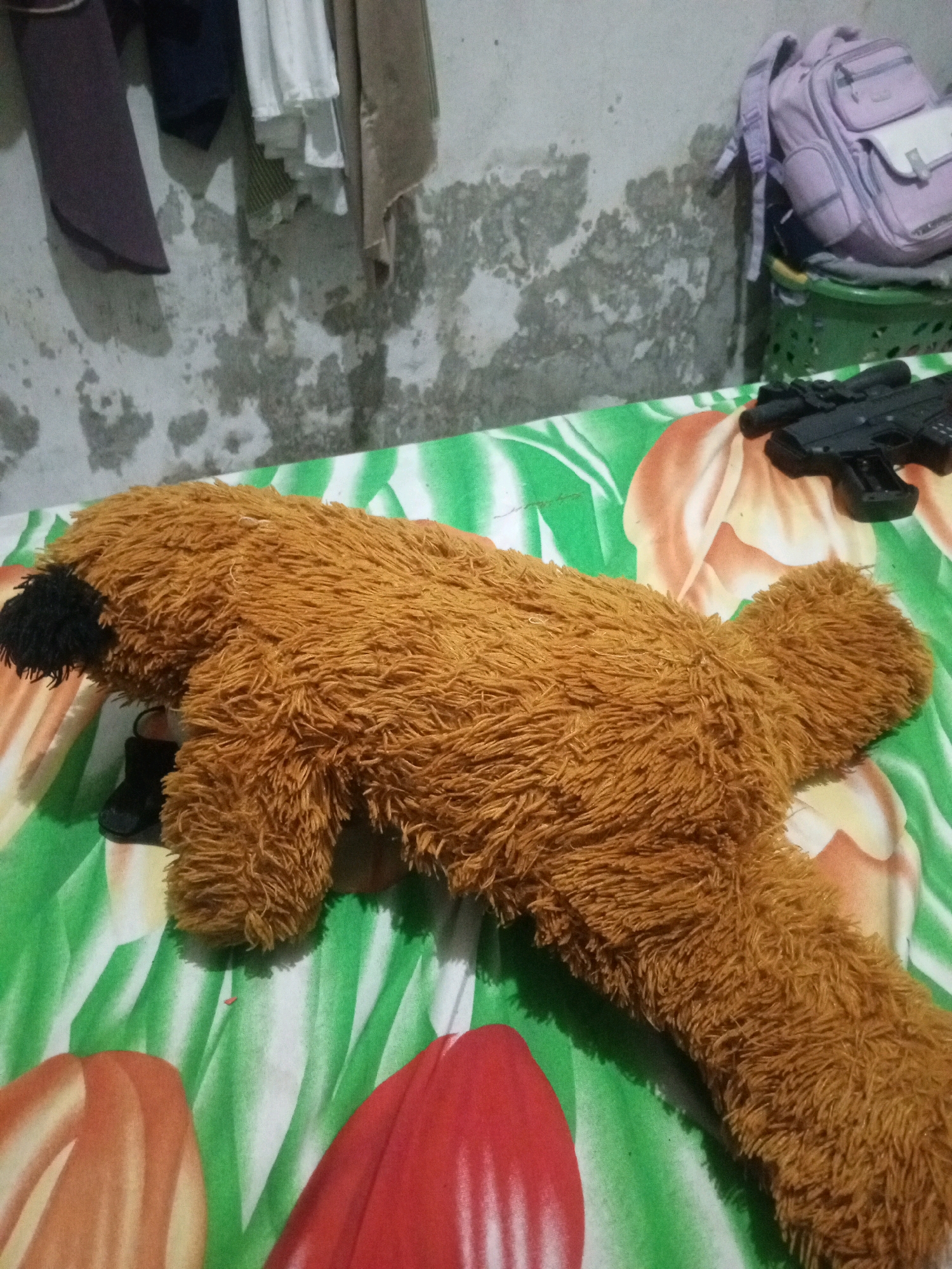 In Kendalpecabean, Indonesia, a brown plush toy dog lies on a patterned tablecloth. The tablecloth features large, abstract shapes in orange, red, and green. To the right of the plush toy, a black toy gun is visible. In the background, a wall with peeling paint is partially obscured by clothing hanging from a bar. A light purple backpack and a green plastic basket are also present in the background.