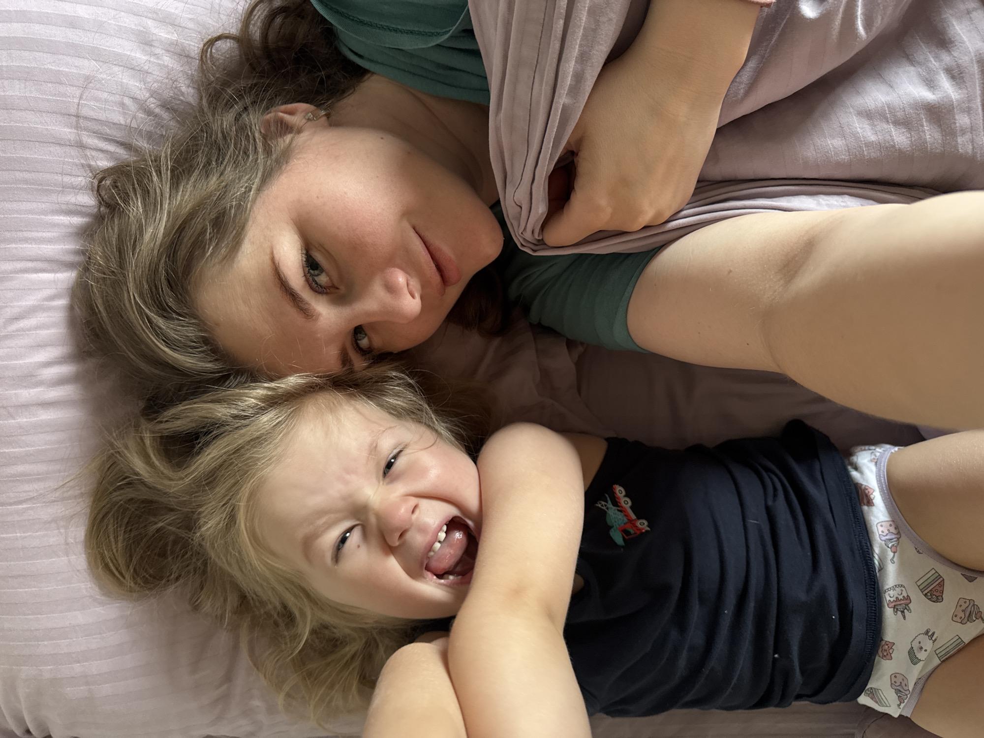 A woman and a child are lying on a lavender-colored bed. The woman, on top, has light brown hair and blue eyes. She is wearing a green t-shirt and holding a lavender-colored fabric. The child, below, has blonde hair and is wearing a navy t-shirt with a graphic of an insect and underwear with illustrations of cupcakes and other sweets. The child is making a distorted expression, with their tongue out and eyes squinted. The photo appears to have been taken from above. The city of Ферма, Russia is not explicitly displayed in this image.