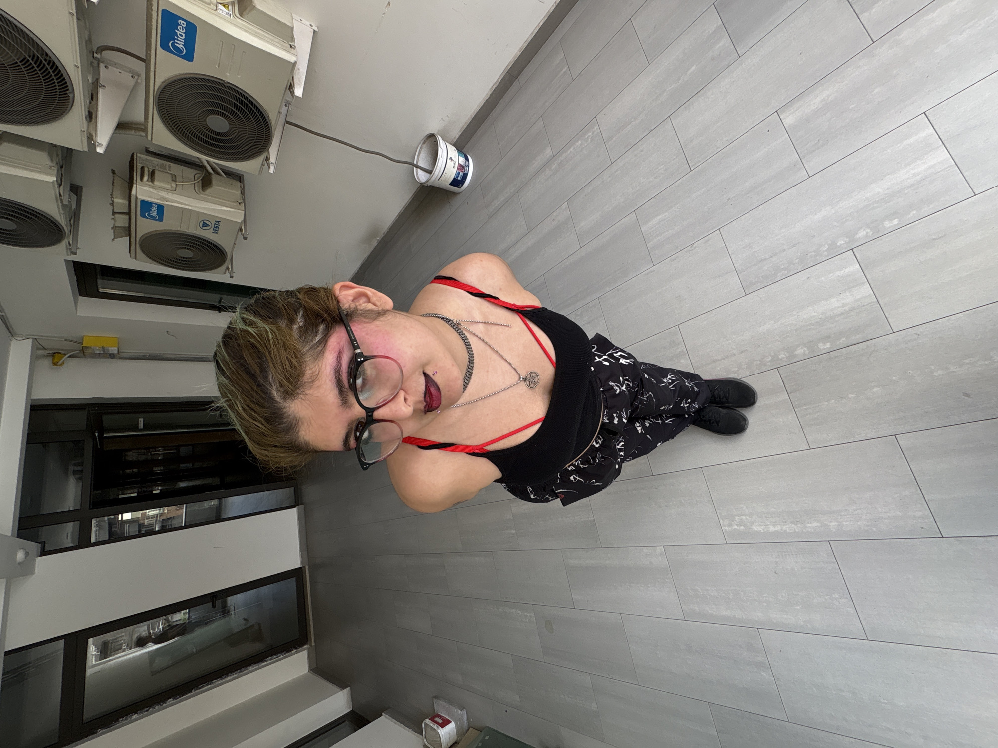 A high-angle, full-body shot captures a young person standing and looking up towards the camera in an indoor or semi-enclosed urban setting in Yerevan, Armenia. The person has light brown hair, possibly with green or blue highlights, and wears large round glasses. They have notable makeup, including dark red lipstick, pinkish eyeshadow, and a piercing above their upper lip. Their attire consists of a black tank top with a visible red strap, a silver chain necklace with a circular pendant, dark pants adorned with a white abstract pattern, and black shoes. Their expression is composed, with a slight smirk as they gaze directly upwards.
The setting features a clean, modern aesthetic with light gray rectangular floor tiles laid in a staggered pattern. Mounted high on a wall or ceiling are several white, outdoor-style air conditioning units, with visible brand names "Midea" and "VESTA." Below these units, a small yellow electrical box is visible on the wall. A white plastic bucket with a wire or hose leading into it rests on the tiled floor near a wall. In the background, dark-tinted windows or glass panels reflect parts of the surroundings, suggesting either an adjacent room or an exterior view. The overall environment appears to be a functional, possibly commercial or public space, well-lit, likely during the daytime. The photograph captures a moment of casual posing.
