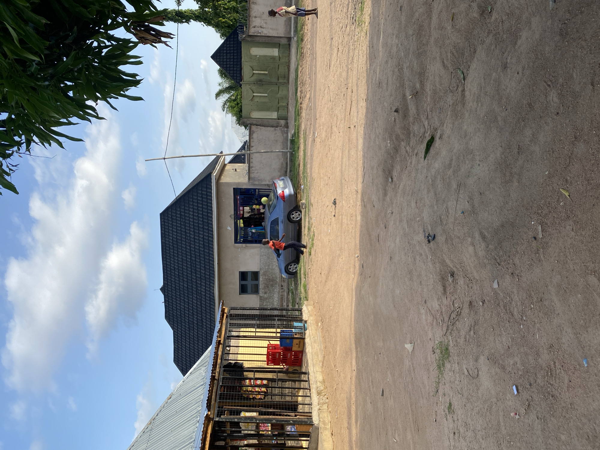 In the city of Howayi, Nigeria, the scene depicts a dusty, unpaved area with several structures and two individuals. To the left, a structure with a corrugated metal roof, resembling a shop or market stall, is enclosed by a metal grate. Inside, various goods are visible, including stacked red crates. Adjacent to this is a building with a dark, tiled roof.
In the center of the frame, a gray sedan is parked. A young boy, wearing an orange shirt and dark pants, is walking towards the right side of the image, near the car. Further to the right, a green metal gate dominates the background, flanked by concrete walls. Another young boy, wearing a light-colored shirt with dark trim and blue shorts, stands facing left near the gate.
The ground is predominantly dirt, with scattered debris and some patches of dry grass. Lush green foliage from a tree hangs into the upper right corner of the image, and a thin wooden pole stands vertically in the middle ground. The sky is blue with scattered white clouds. The lighting suggests it is daytime, likely late afternoon or early evening due to the shadows.