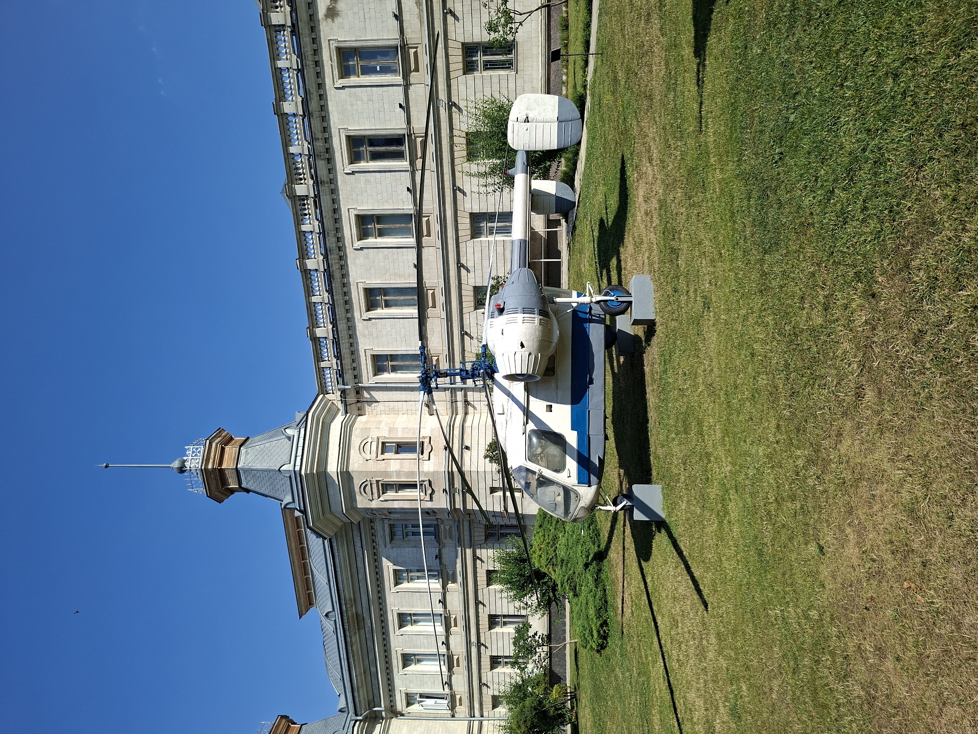 The image shows a light-colored helicopter with blue accents parked on a grassy lawn in front of a light-colored building. The building has a classic architectural style with multiple windows and ornate detailing along the roofline. A prominent tower with a pointed roof and a spire extends from the building. The sky is clear and blue, suggesting a sunny day, likely daytime given the bright lighting and strong shadows cast by the helicopter. The grass is well-maintained but shows some dry patches. The helicopter appears to be on display or static exhibit rather than in active flight. There is no visible text in the image. The scene evokes a sense of stillness and perhaps historical significance, with the helicopter juxtaposed against the traditional architecture.