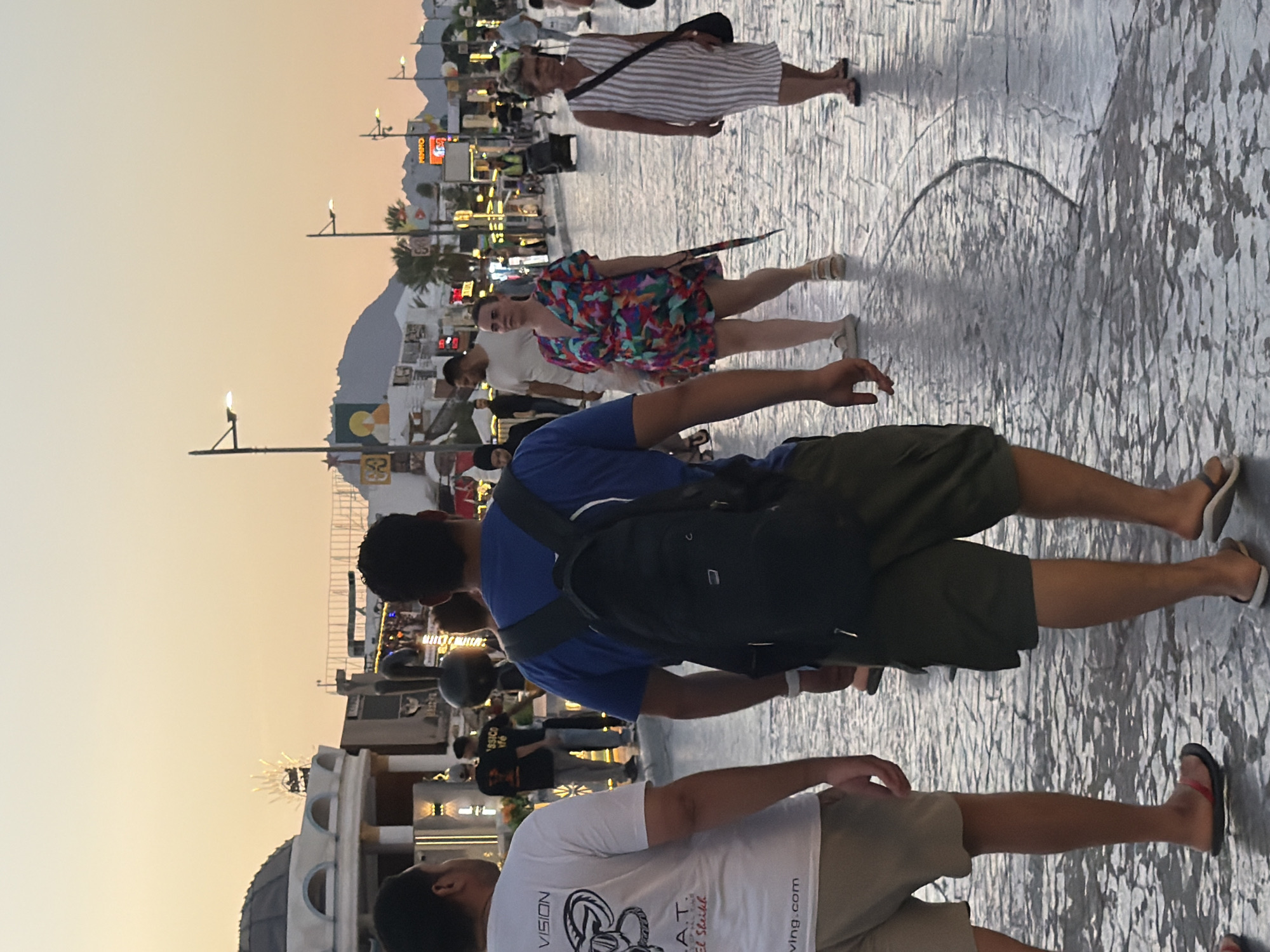 In this vertical, eye-level, full-shot image, a group of tourists stroll along a paved walkway in Sharm El Sheikh, Egypt, at dusk. The sky is a gradient of pale yellow and orange, suggesting the sun is setting. In the foreground, a man wearing a white t-shirt with graphic print and tan shorts walks away from the camera. To his right, another man in a blue t-shirt and olive-green shorts, carrying a black backpack, also walks away. Further back, a woman in a colorful, abstract-print romper and a woman in a white and beige striped dress walk side-by-side, also moving away from the camera. In the background, more people are visible walking, as well as several shops with illuminated signs. The architecture appears to be of a resort town, with dome-like structures and brightly lit facades. The paved walkway is textured and appears wet in places, reflecting the ambient light. Several lampposts are visible along the sides of the walkway.