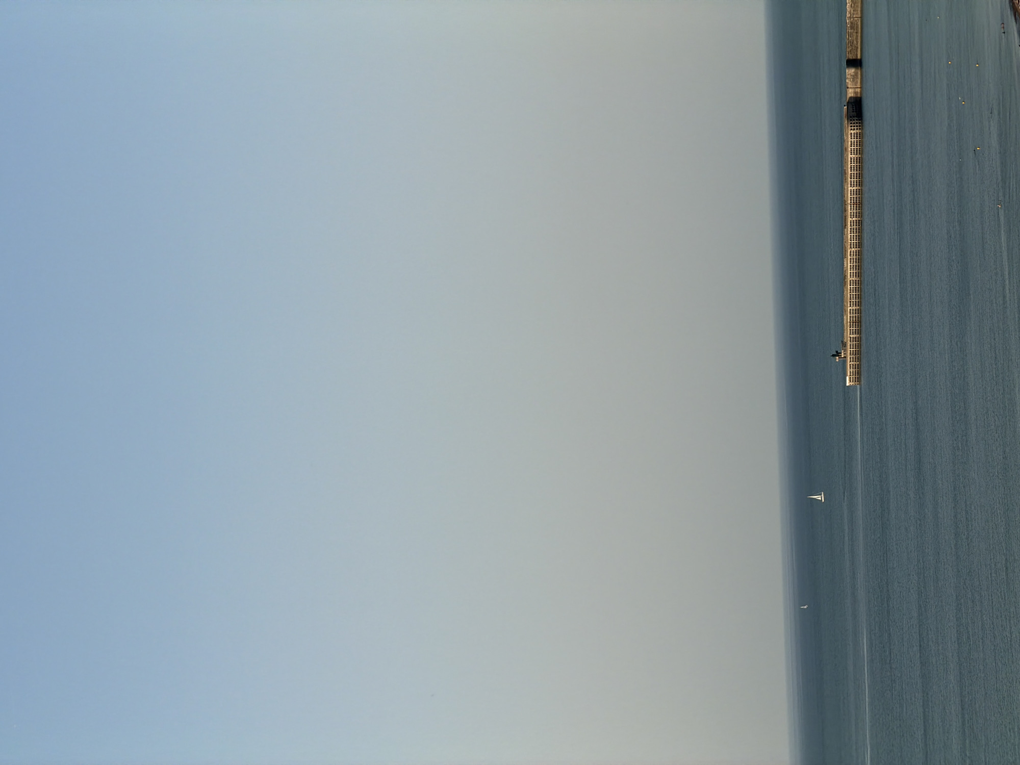 This is an aerial, wide shot of a calm, blue sea with a long pier extending into the water from the right side of the frame. The pier has a weathered, wooden surface and appears to be a popular spot for recreation, with several small yellow buoys floating in the water near it. In the distance, two sailboats are visible on the water, one larger and closer than the other. The sky is a pale, hazy blue, suggesting it might be early morning or late afternoon, with the sun not directly visible. The overall atmosphere is peaceful and serene. There are no people or visible text in the image. The image was taken in Dieppe, France.