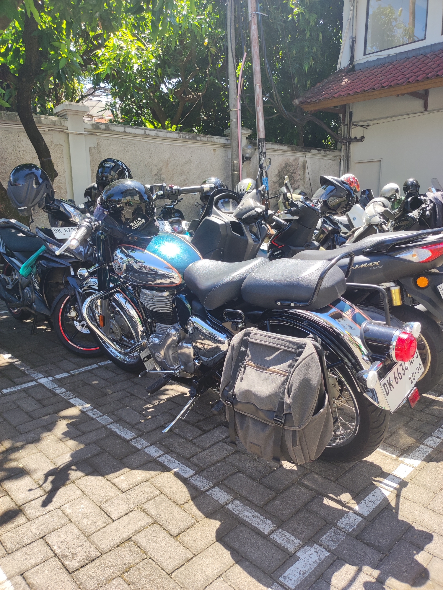 The image shows a parking area filled with motorcycles in Denpasar, Indonesia. The motorcycles are parked in marked spaces on a brick pavement surface. Sunlight casts shadows across the scene. The motorcycles are of various models and colors, including black, blue, and chrome. A helmet rests on each motorcycle. A gray saddlebag is attached to the rear of one motorcycle. A white building with a red tile roof is in the background. Trees are also visible in the background.