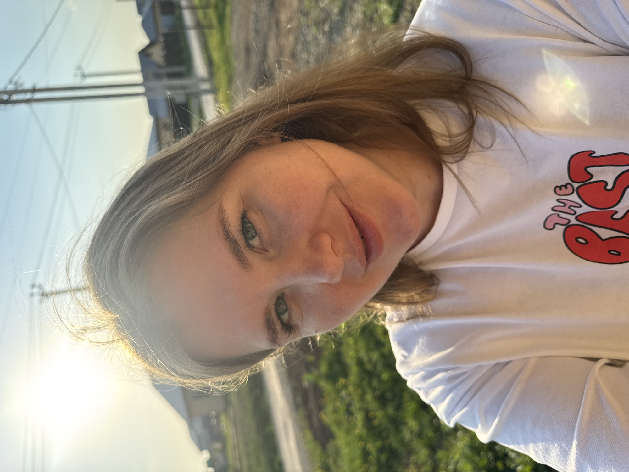 A young woman with light brown hair and fair skin smiles at the camera. She has green eyes and freckles are visible on her face. She is wearing a white t-shirt with red lettering on the chest.
The background is blurred but shows greenery and possibly a street, and building in the far distance. The sunlight is bright, causing lens flare. The image is taken outdoors, possibly in Ферма, Russia, given the location details.