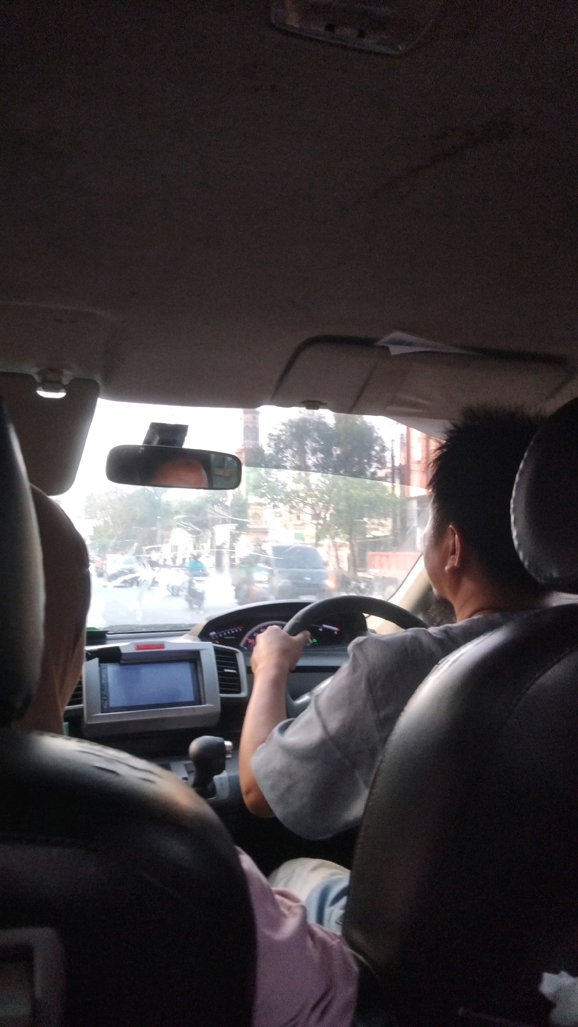 The image captures an interior view of a car, likely in Samarinda, Indonesia, based on contextual information. A male driver is visible from the shoulders up, operating the steering wheel of the vehicle. He is wearing a grey t-shirt. The car's dashboard features a car stereo system with a screen, and a gear shift lever is also discernible. Outside the car, through the windshield, a street scene unfolds with other vehicles and buildings suggesting an urban environment. The lighting indicates it is daytime, possibly late afternoon or early morning due to the softer light. The view from inside the car is slightly obstructed by the car's interior elements like the rearview mirror and the car's headliner.