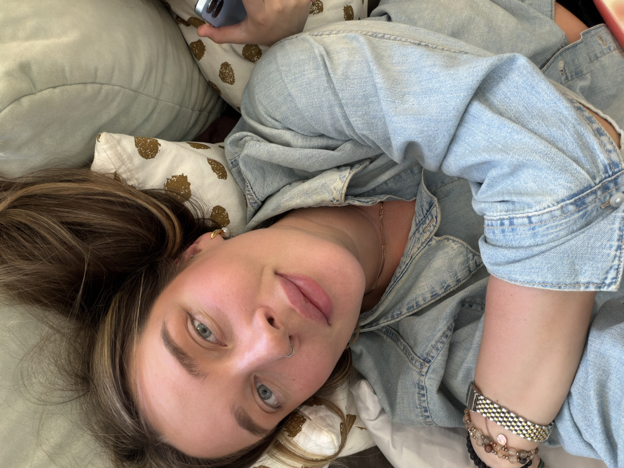 A woman is lying on a pillow, likely indoors, wearing a light-wash denim jacket. She is looking at the camera, holding a blue cell phone with the camera facing towards her head. The location is unknown, and there's no information to suggest it is Chaikovsky, Russia.